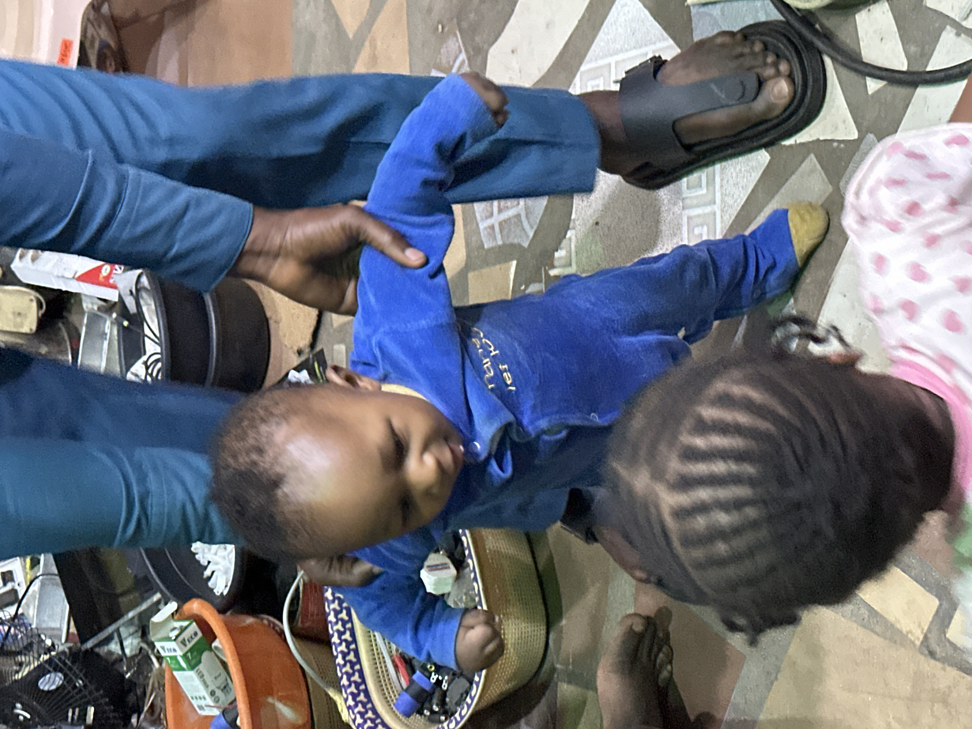 The image depicts an indoor scene in Narayi, Nigeria, featuring three individuals: an adult and two children. In the center, a baby, dressed in a royal blue, full-body outfit with yellow-colored foot soles, stands upright, supported by the hands of an adult. The adult, whose lower torso and legs are visible, wears teal trousers and a dark, open-toed sandal on the right foot. The adult's dark-skinned hands are positioned at the baby's upper arms/shoulders. In the immediate foreground, with their back to the viewer, is a child with dark hair styled into distinct linear patterns on the scalp, wearing a light-colored garment with a patterned print.
The flooring consists of light-colored tiles with a repeating geometric pattern of darker lines forming squares and crosses. To the mid-left, a light-colored woven basket with a patterned rim holds an assortment of small, dark objects, a blue roll of tape, and a small white rectangular object. Further left, an orange plastic basin contains a white box with green markings (partially visible "VECO") and other items. A portion of a small electric fan is visible in the upper-left background, alongside what appears to be a power outlet. The background elements suggest a functional or storage area with some visual clutter.