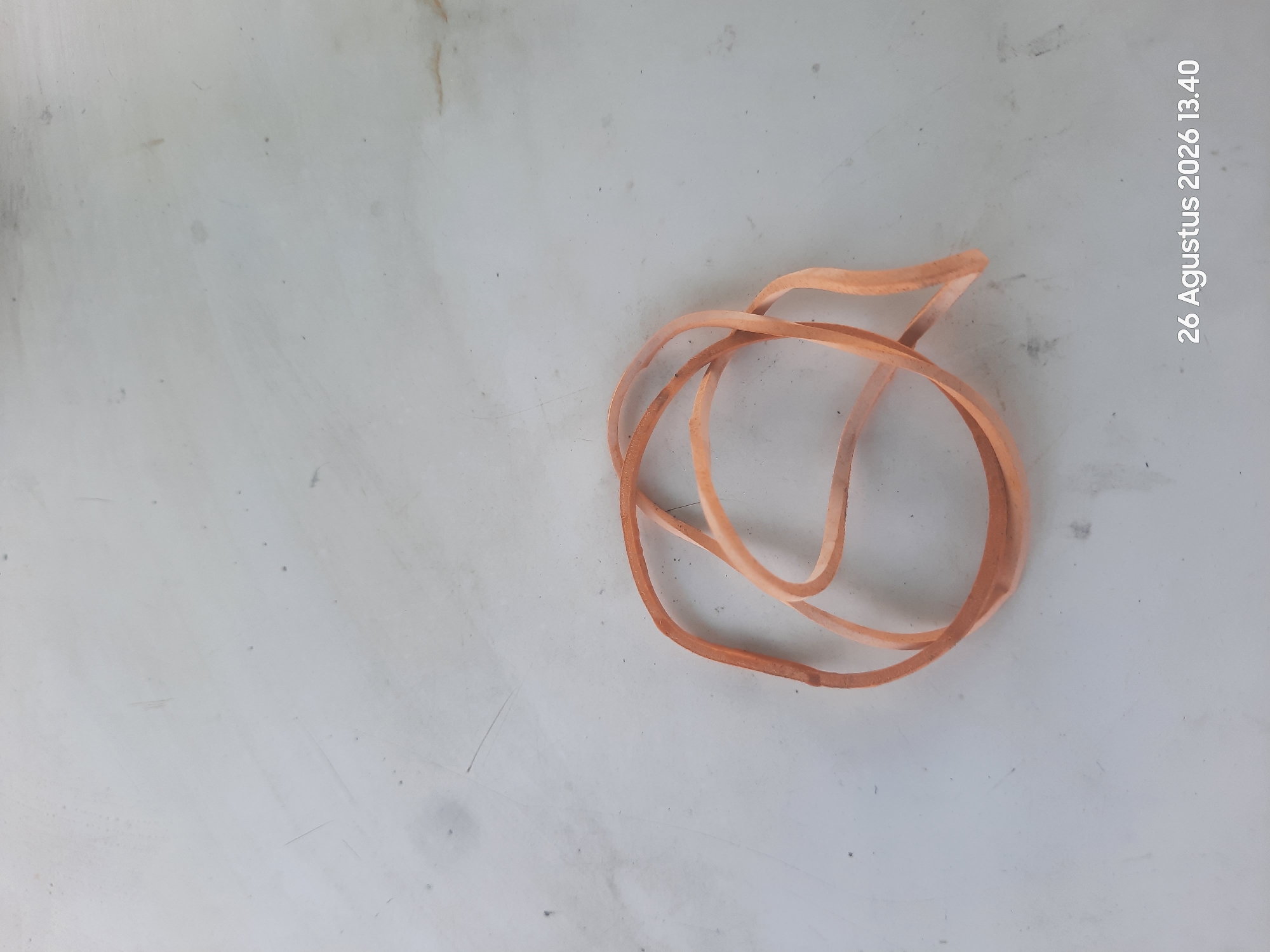 An overhead, medium-shot photograph shows a close-up of a tangled bundle of tan-colored rubber bands lying on a light-colored, textured surface. The rubber bands are circular and appear to be a standard size. The surface has faint scratches and smudges, suggesting it may be a table or countertop. The upper portion of the image is primarily empty space, with the rubber bands occupying the lower-middle section. The bottom right corner of the image displays a timestamp: "26 Agustus 2026 13.40". The scene is indoors and appears to be a simple, static arrangement. There are no people or animals present, and no discernible activity is taking place. The lighting is even, with no harsh shadows. The location is identified as Gending, Indonesia.