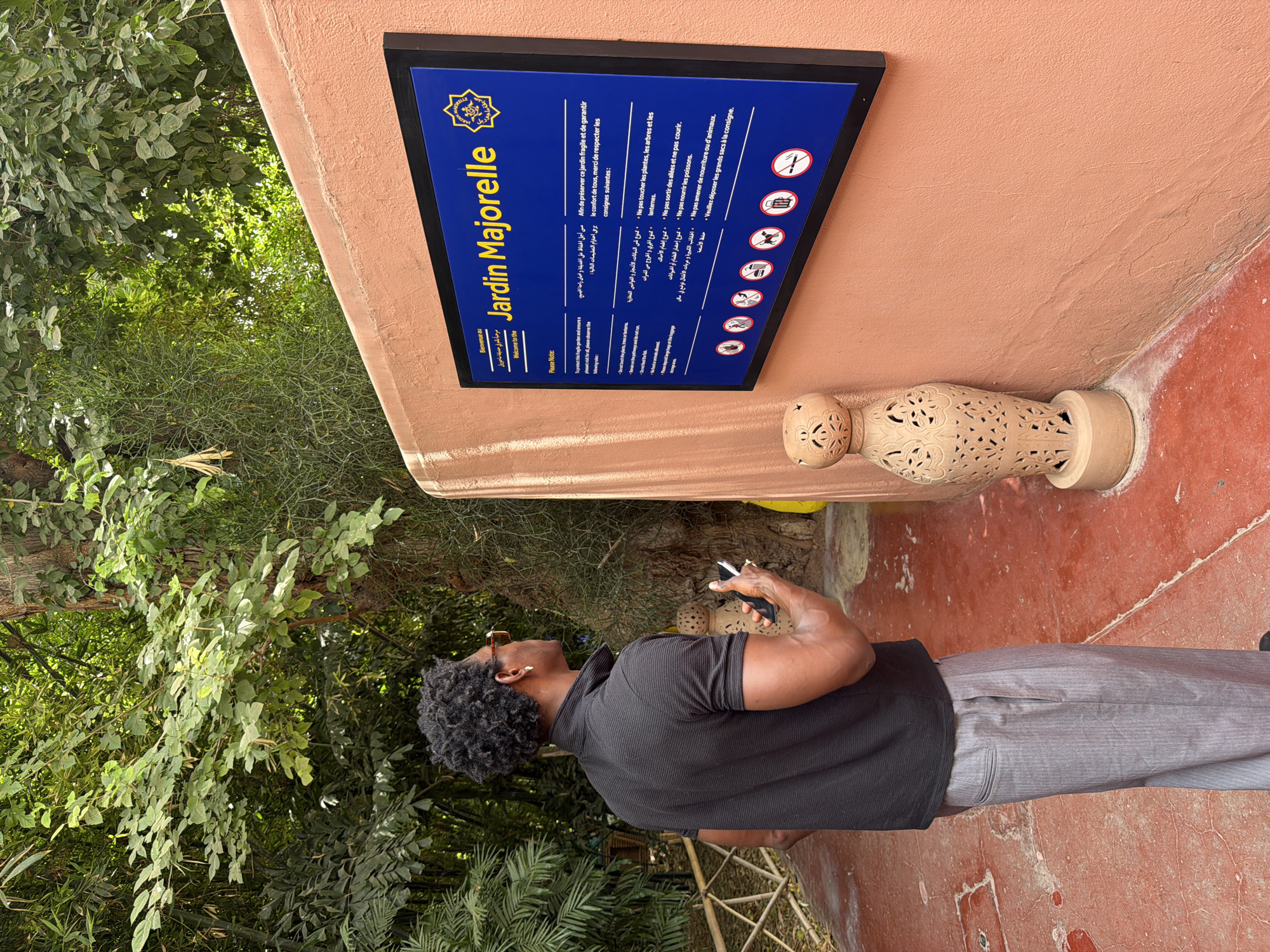 This media file features a man standing in front of a sign at the Jardin Majorelle in Marrakesh, Morocco. The sign, displayed on an orange wall, is predominantly blue with yellow text and a decorative crest. It welcomes visitors and outlines rules for preserving the garden, with text in French, Arabic, and English. The rules, accompanied by pictograms, prohibit touching plants, running, feeding fish, and bringing food or animals. Large bags are to be left at a locker.
The man, wearing a black polo shirt and grey trousers, is looking upwards towards the sign, holding a smartphone in his right hand. His attention is directed at the information presented. The surrounding environment is lush with green foliage and trees, typical of a botanical garden. The ground appears to be a reddish-orange, textured surface. The lighting suggests it is daytime, with sunlight filtering through the trees, creating dappled light and shadow. The overall scene conveys a sense of respectful observation of the garden's rules and an appreciation for its natural beauty. A decorative ceramic object is mounted on the wall near the sign, adding an artistic element to the setting.