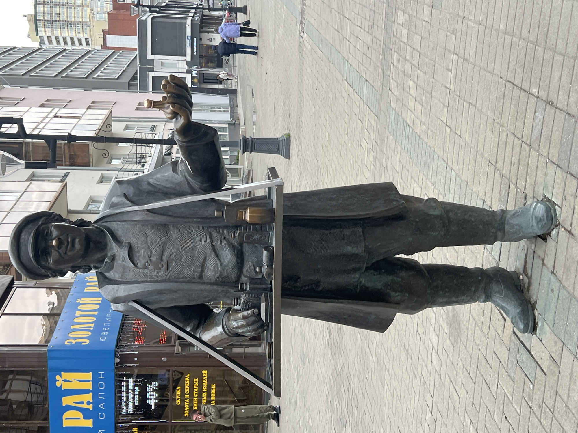 The image shows a bronze statue of a street vendor in Yekaterinburg, Russia. The statue is located on a pedestrian street in front of a storefront with the words "ЗОЛОТОЙ РАЙ" (Golden Paradise) and "ЮВЕЛИРНЫЙ САЛОН" (Jewelry Salon) visible on a blue sign. The vendor statue is holding a tray with small bottles and containers. A person wearing a green tracksuit is standing to the left of the statue, on their phone. In the background, several people are walking down the street, and there are buildings on either side of the street. The ground is paved with brick-like tiles.