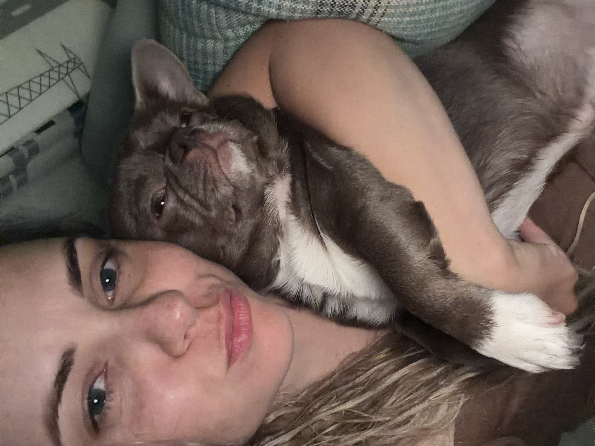 A woman lies on a bed, holding a brown and white French Bulldog. The location cannot be determined as Votkinsk, Russia. The woman has fair skin, light blonde hair, and blue eyes. Her facial expression is relaxed. The dog is nestled on her chest and arm. The background shows a bed with a patterned pillow.