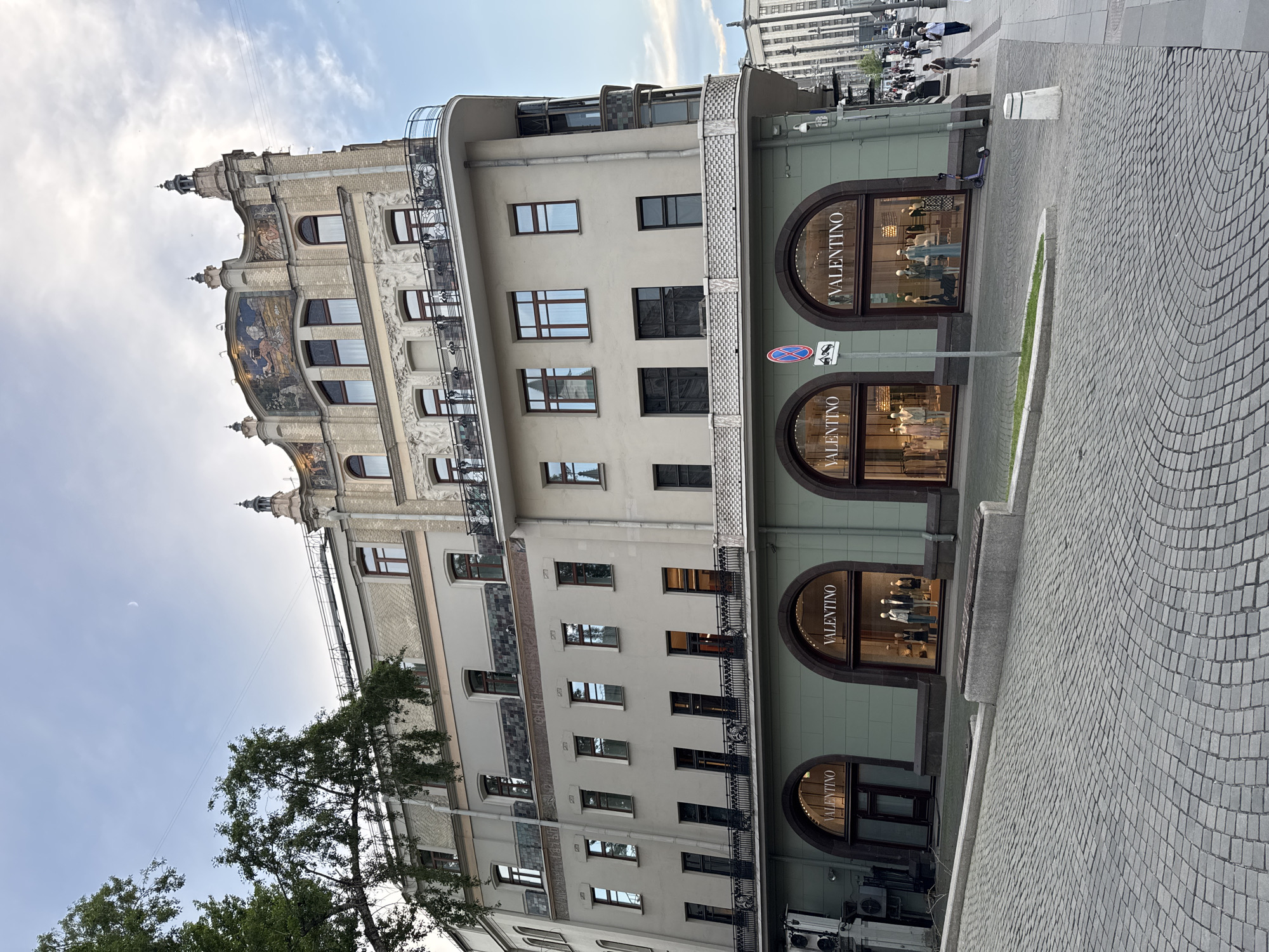 The image shows a multi-story building in Moscow, Russia, featuring a Valentino store at street level with four arched window displays. The building's architecture combines classical elements with modern design. A visible sign prohibits bicycles, and there is pedestrian activity on the sidewalk in the background. The street is paved with cobblestones.