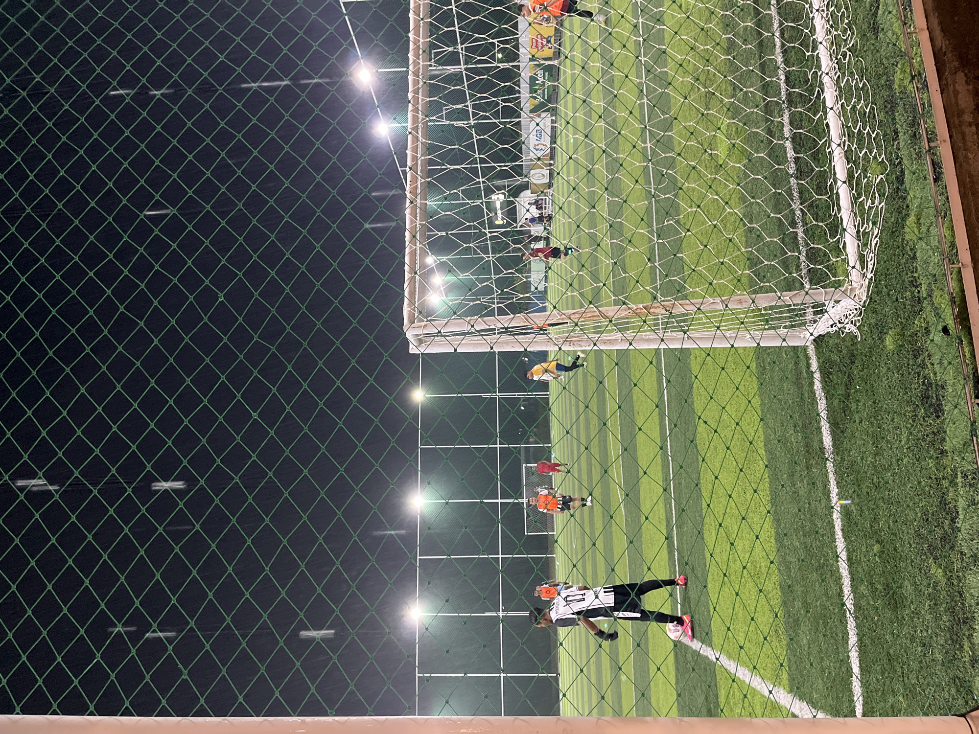 The image shows a soccer game in progress, viewed through a chain-link fence and a soccer goal net. Several players are visible on the green artificial turf field under bright artificial lights. One player in a black and white jersey and black shorts is positioned near the bottom of the image, appearing to prepare for a kick. Other players, some wearing orange jerseys, are scattered across the field. In the background, behind the goal, is a banner with text and logos. The scene appears to be located in Lhokseumawe, Indonesia, evidenced by the text "BANK ACEH" visible on the banner in the background. The time appears to be night due to the darkness surrounding the field and the use of artificial lighting.