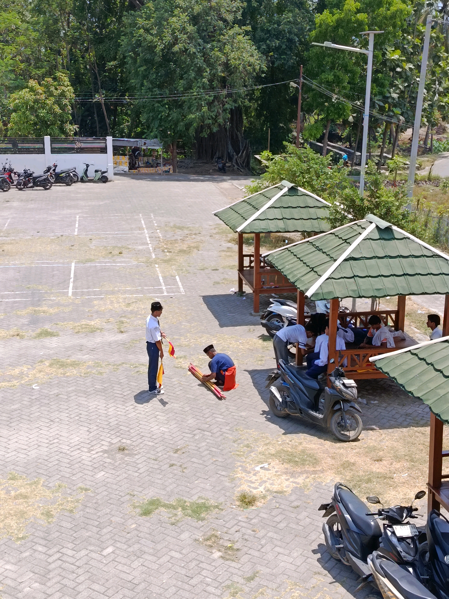 The image shows an outdoor scene in Ramban Wetan, Indonesia. In the foreground, two young males are present on a paved area with a pattern of interlocking gray bricks. One, wearing a white shirt, dark trousers, and a black cap, stands holding two flags, one red and one yellow. The other male, wearing a blue shirt and orange shorts, kneels on the ground and gathers a stack of red and yellow cylindrical objects, possibly flags or markers, with his hands. Nearby, parked on the brick pavement, are several motorbikes, including a dark-colored motorbike in the lower right and a lighter-colored motorbike to its left.
In the mid-ground, under two green-roofed wooden gazebos, several other young males are seated or standing. The gazebos have open sides and wooden supports. One gazebo has a white motorbike parked next to it. In the background, a white fence runs along the edge of the scene, with parked motorcycles behind it. Beyond the fence, a dense area of trees forms a natural backdrop, with some electrical poles and wires visible. The ground in the open areas appears to be a mix of paved brick and dry, patchy grass. The lighting suggests it is daytime, with distinct shadows cast by the people and structures.