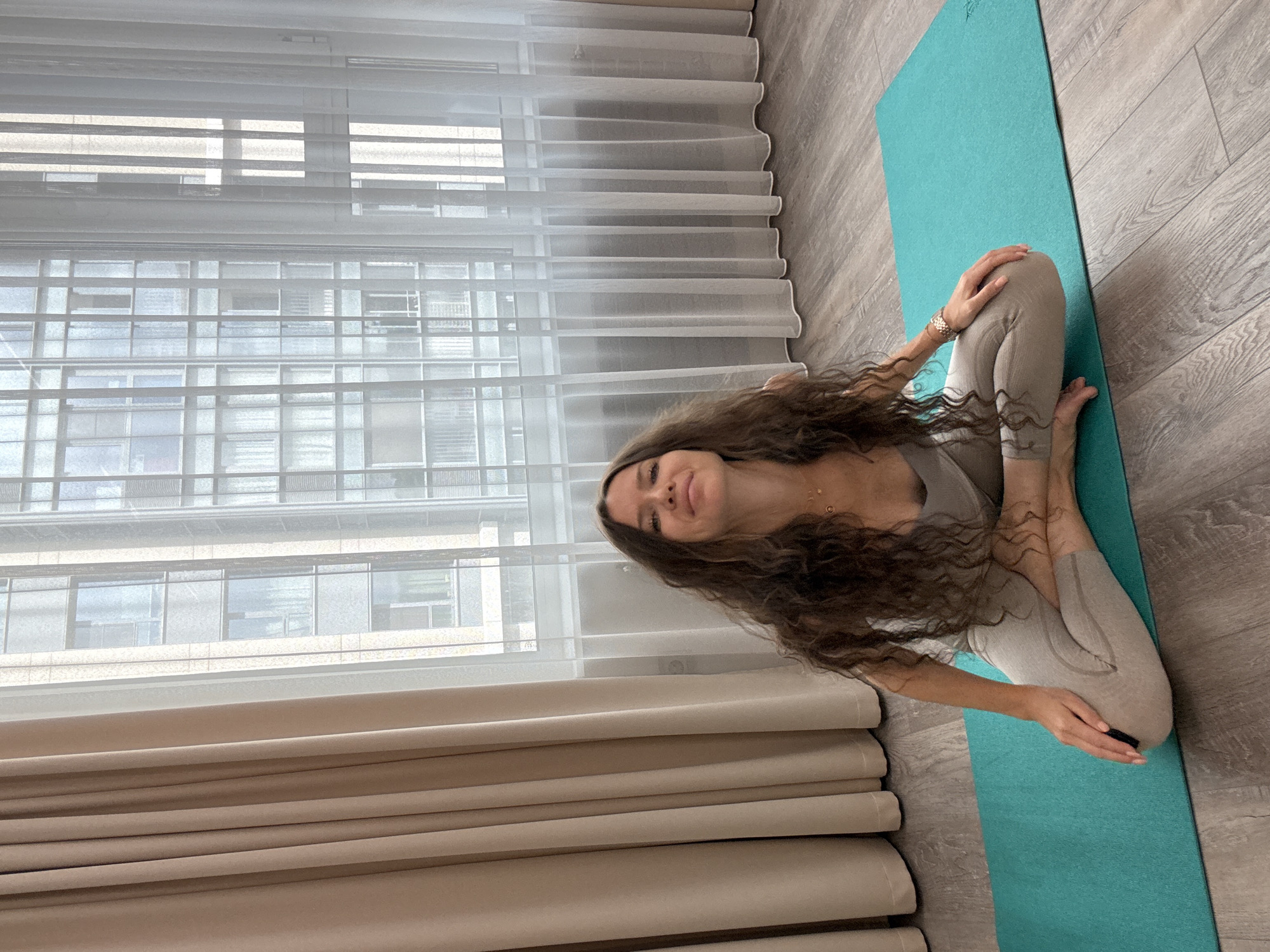 A woman with long, curly brown hair is sitting cross-legged on a teal yoga mat on a wooden floor. She is wearing a light grey tank top and light grey leggings. She is smiling and looking at the camera. Behind her, there are large windows covered by sheer white curtains and heavier beige curtains. The windows overlook a city building. The scene appears to be an indoor space, possibly a living room or a yoga studio, in the city of Khimki, Russia.