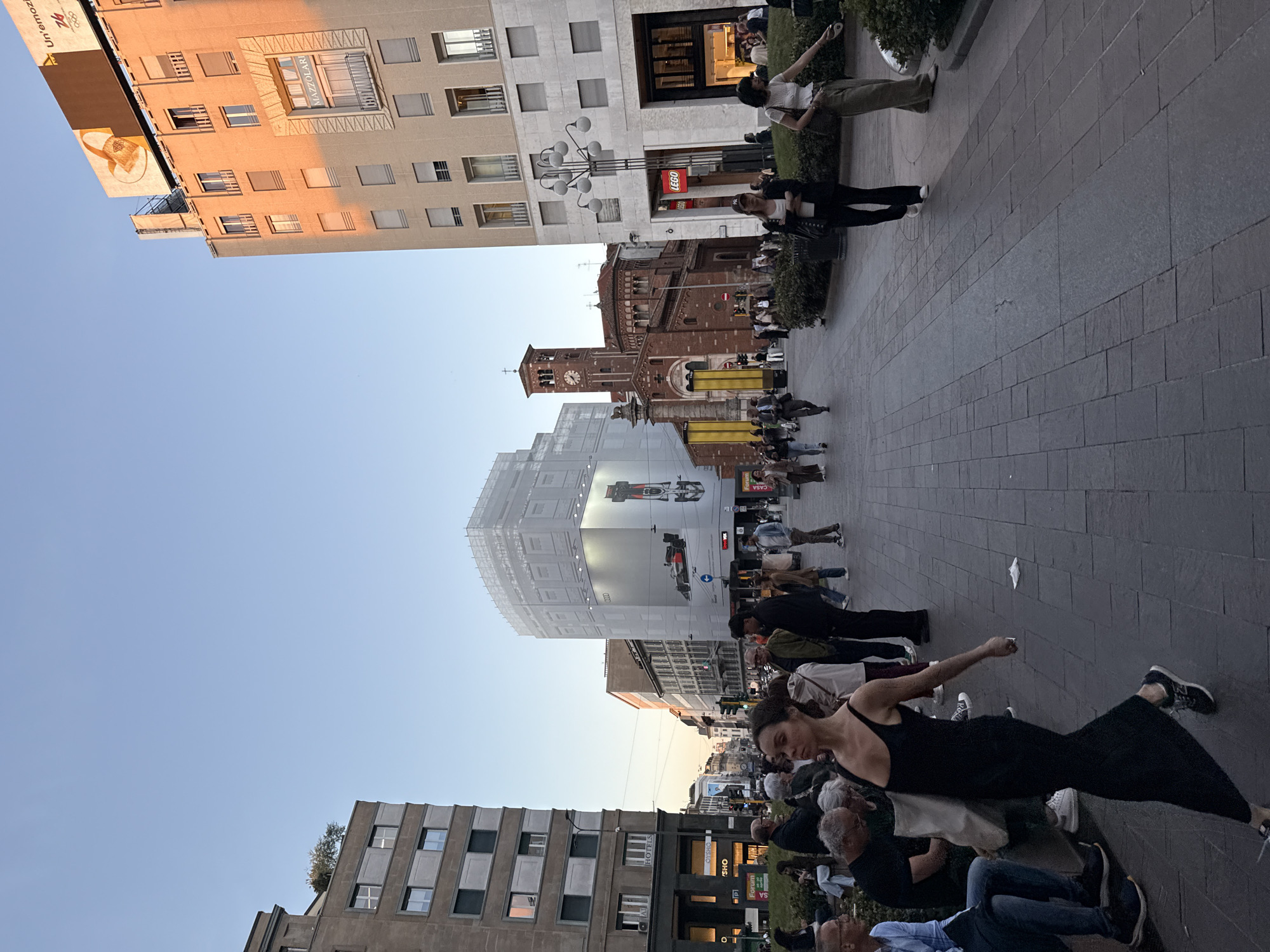 The image, rotated 90 degrees clockwise, captures a lively urban scene in what appears to be a city square or broad street, likely in Milan, under a clear blue sky during the late afternoon or early evening. Warm, golden sunlight illuminates the facades of buildings on the left side of the frame.
In the foreground, a woman wearing a black sleeveless dress and dark sneakers stands on a grey paved surface, her left arm outstretched in a pose that suggests dancing or performing. Several other individuals are seated or standing around her, some visible only partially.
The midground features a mix of architectural styles. To the right, a beige building houses a "LEGO" store at street level, beneath a distinctive multi-bulb street lamp. Further down, a historical red-brick church with a prominent clock tower and spires stands majestically, indicating an older part of the city. To the left, a contemporary building features a large, reflective surface displaying a billboard with an "AUDI" branded race car. People are seen walking, standing, and sitting at various outdoor establishments along the street.
In the background, towering commercial or residential buildings line the street. One building has the name "MAZZOLARI" inscribed on an ornate panel, and a larger billboard above it shows partially visible text including "Un'emozio..." and "2026," potentially referencing the Milan Cortina Olympic Games. Other visible text elements include "HOTELS," "OSHO," and "Forum P1." The overall impression is one of a bustling city center with a blend of historical and modern elements, filled with people enjoying the urban environment.