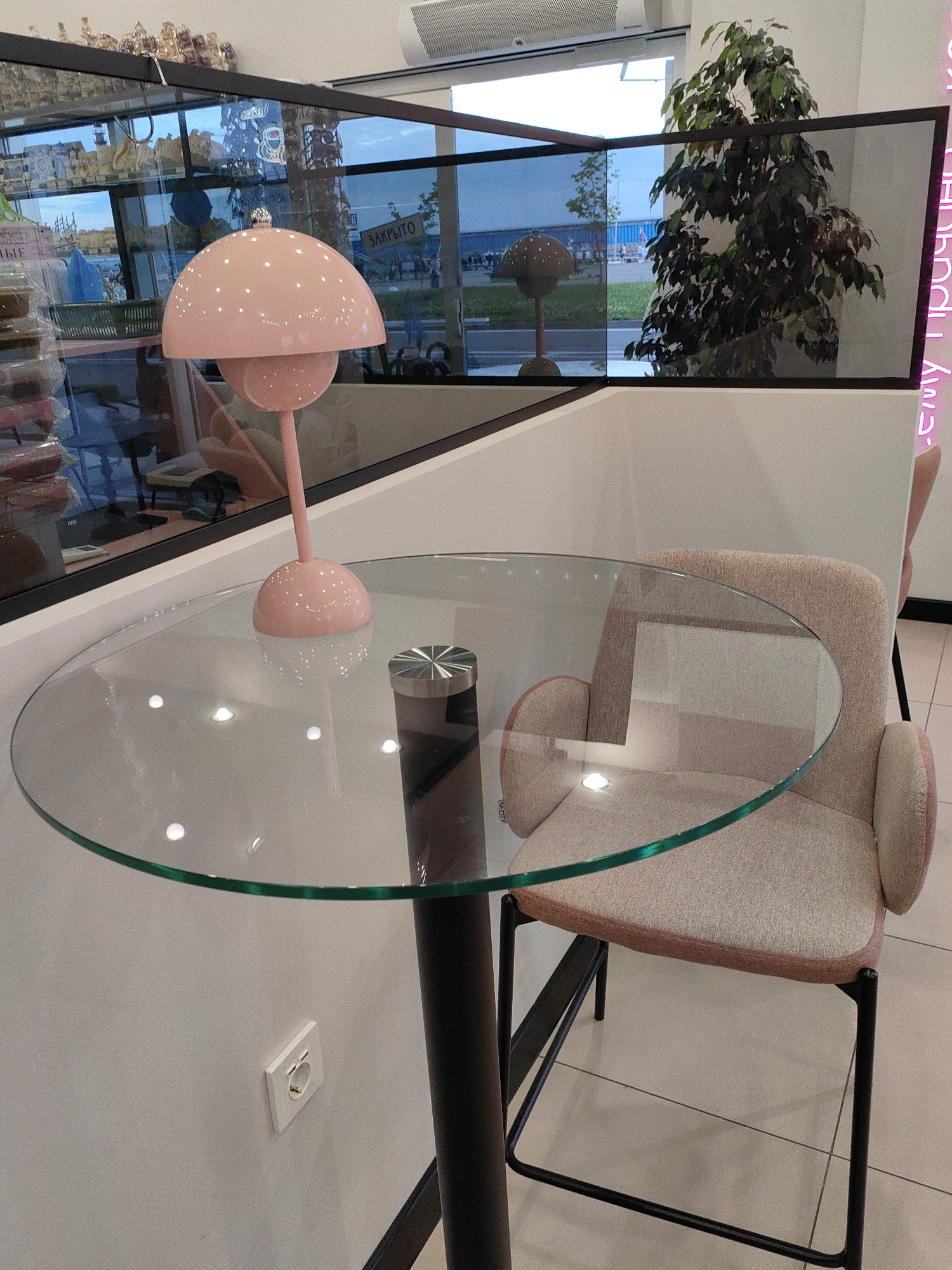 This media file depicts a modern interior space, likely a cafe, lounge, or retail area in Sirius, Russia, furnished with a glass-top table and a high chair. The scene is bright and clean, characterized by light colors and contemporary design elements.
In the foreground, a round glass table with a dark, cylindrical base stands centrally. On the table, a distinctive pink mushroom-shaped lamp with a glossy finish and small reflective dots serves as a decorative element. Adjacent to the table is a comfortable-looking high chair or bar stool, upholstered in a light beige or pale pink fabric, featuring a sleek black metal frame. A white electrical outlet is visible on the wall beneath the table.
The background is dominated by a large glass partition that separates the interior from the outside. Through this glass, an outdoor scene is visible, featuring a clear blue sky, distant structures, and possibly a street or open area. Another pink mushroom-shaped lamp, identical to the one on the table, appears either reflected in the glass or positioned outside as part of an exterior display. A tall, lush potted plant with dark green leaves, possibly a Ficus, stands behind the glass partition. On the left side, behind the glass, other retail displays or products can be discerned.
The setting appears to be during daytime, as indicated by the natural light illuminating the exterior. No people are visible, suggesting the space might be empty, closed, or captured during off-peak hours.
Notable text includes a sign visible through the glass windows displaying the Russian word "ЗАКРЫТО" (ZAKRYTO), which translates to "CLOSED". On the right side of the image, part of a neon sign in Russian is visible, reading "Ему приче..." (Emu priche...), which is incomplete but suggests a word like "прическа" (hairstyle) or a phrase with "причинено" (caused). The overall atmosphere conveys a sense of calm and modern aesthetics.