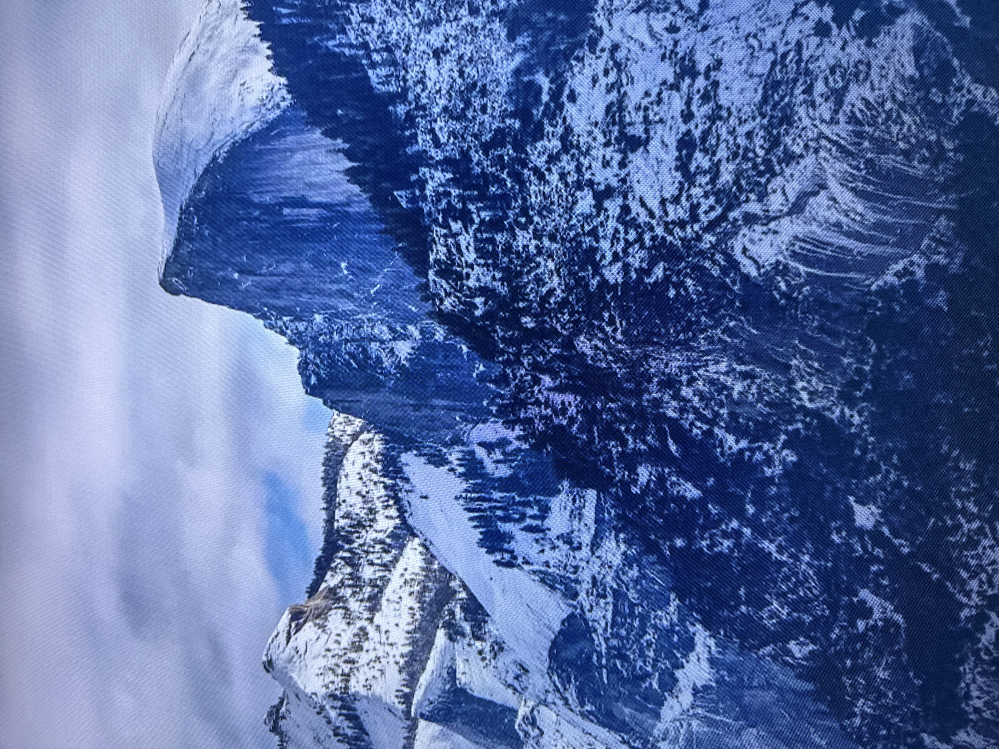The image displays a majestic snow-covered mountain range under a cloudy sky. The dominant feature is a large, sheer cliff face, partially covered in snow, rising sharply to a rounded peak. To the left, another snow-capped mountain with a jagged summit is visible, interspersed with dark evergreen trees. The lower slopes of the mountains are densely forested with snow-laden pines, creating a stark contrast between the white snow and the dark green foliage. The sky is filled with a mix of white and grey clouds, with a small patch of blue sky visible in the distance. The overall impression is one of a rugged and wild natural landscape in winter. There is no indication of the city of Edmonton, Canada in this image.