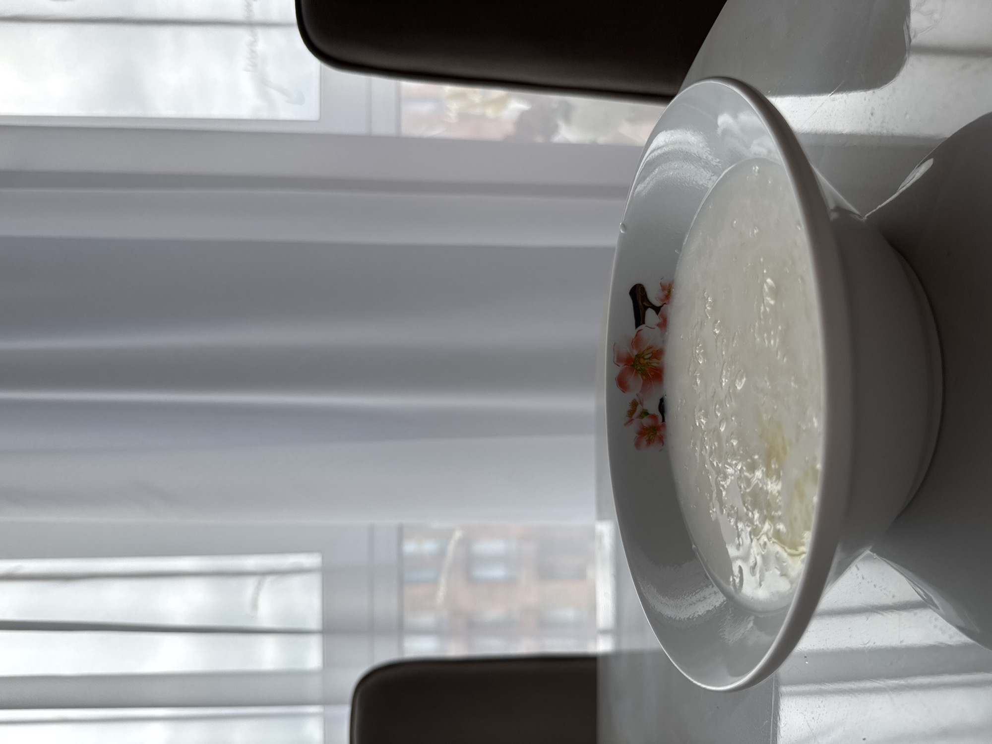 A close-up, low-angle shot shows a white bowl of white porridge on a reflective white table. The bowl has a floral pattern with pink and orange flowers and green leaves on its rim. The porridge appears to have some butter or oil on top. In the background, through sheer white curtains, there are hints of buildings and an overcast sky, suggesting an indoor setting in Laikovo, Russia. Two dark-colored chairs are partially visible on either side of the table, framing the bowl of porridge. The lighting is soft and diffused, coming from the window.