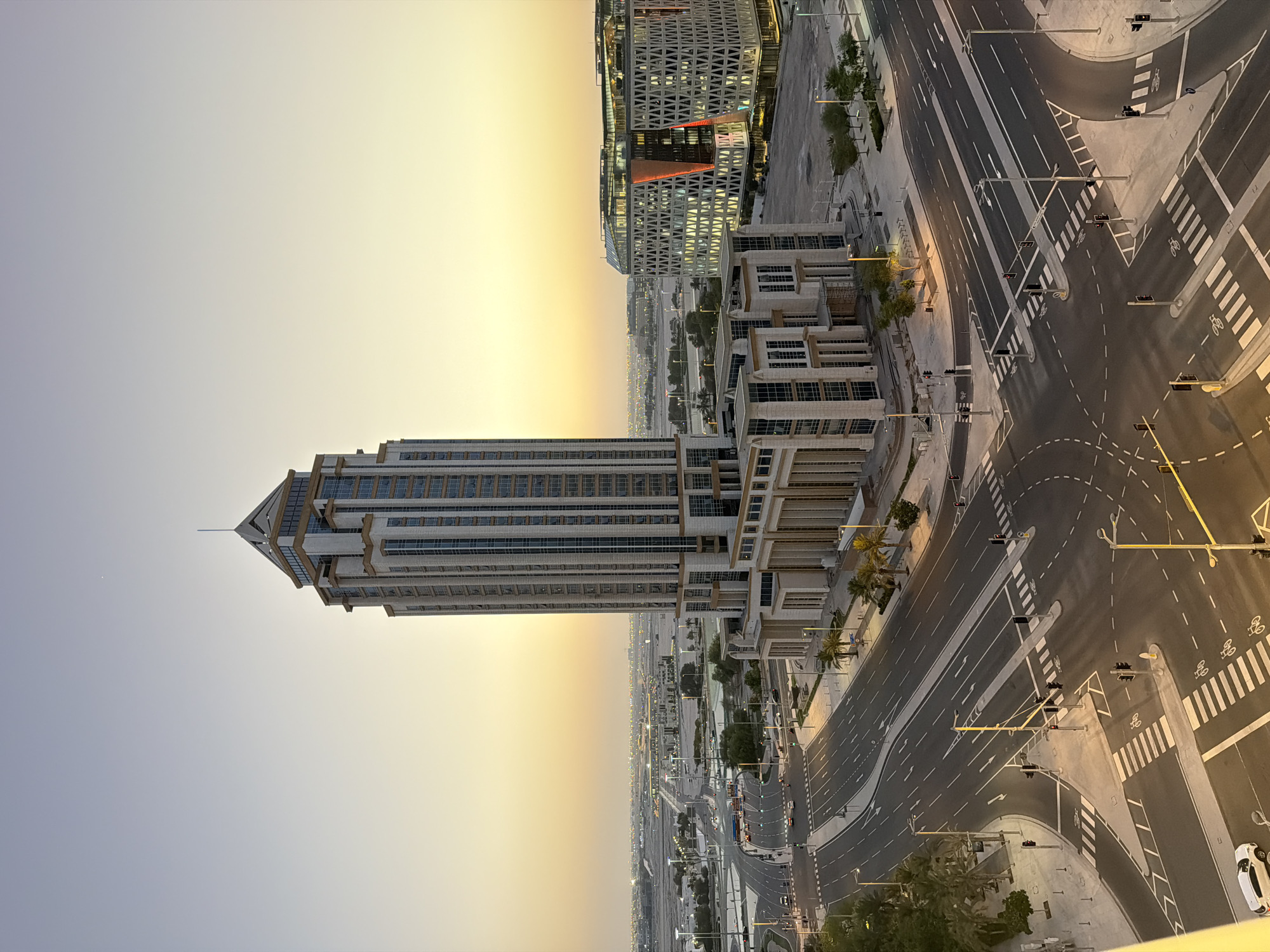 The image depicts an urban landscape at dusk or dawn, characterized by a gradient sky transitioning from a pale blue to a warm yellow-orange near the horizon. Dominating the foreground and midground is a tall, modern skyscraper with a light-colored facade and numerous rectangular windows. To the right of this skyscraper, a contemporary building with a geometric, patterned facade and illuminated windows is visible. The lower portions of the buildings feature classical architectural elements like columns. The scene is set against a backdrop of a sprawling city with distant lights and structures, suggesting a large metropolitan area. In the foreground, a multi-lane intersection with clear lane markings and pedestrian crossings is present. Several traffic lights are visible, some indicating red. A white car is parked on the far left. The roads are paved and well-maintained, with some trees and streetlights lining the sides. The overall impression is one of a modern, developed urban environment.