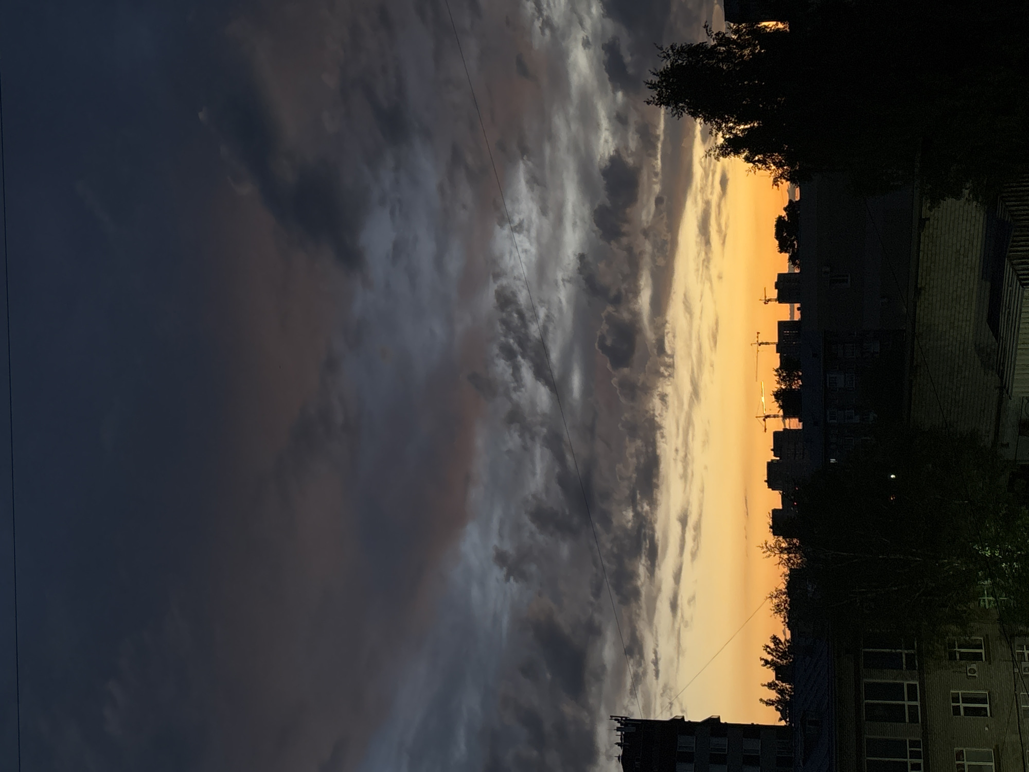 The image captures an evening urban scene. Dark, heavy clouds dominate the left portion of the frame, transitioning to a vibrant orange-yellow sunset sky on the right. The silhouettes of buildings and trees are visible against the bright sky, interspersed with what appear to be construction cranes. Residential buildings with windows and visible details are located on the right, near the bottom corner. A single power line stretches across the sky. The general location appears to be Perm, Russia based on the urban landscape with residential buildings, trees, and power lines, under a cloudy sky.