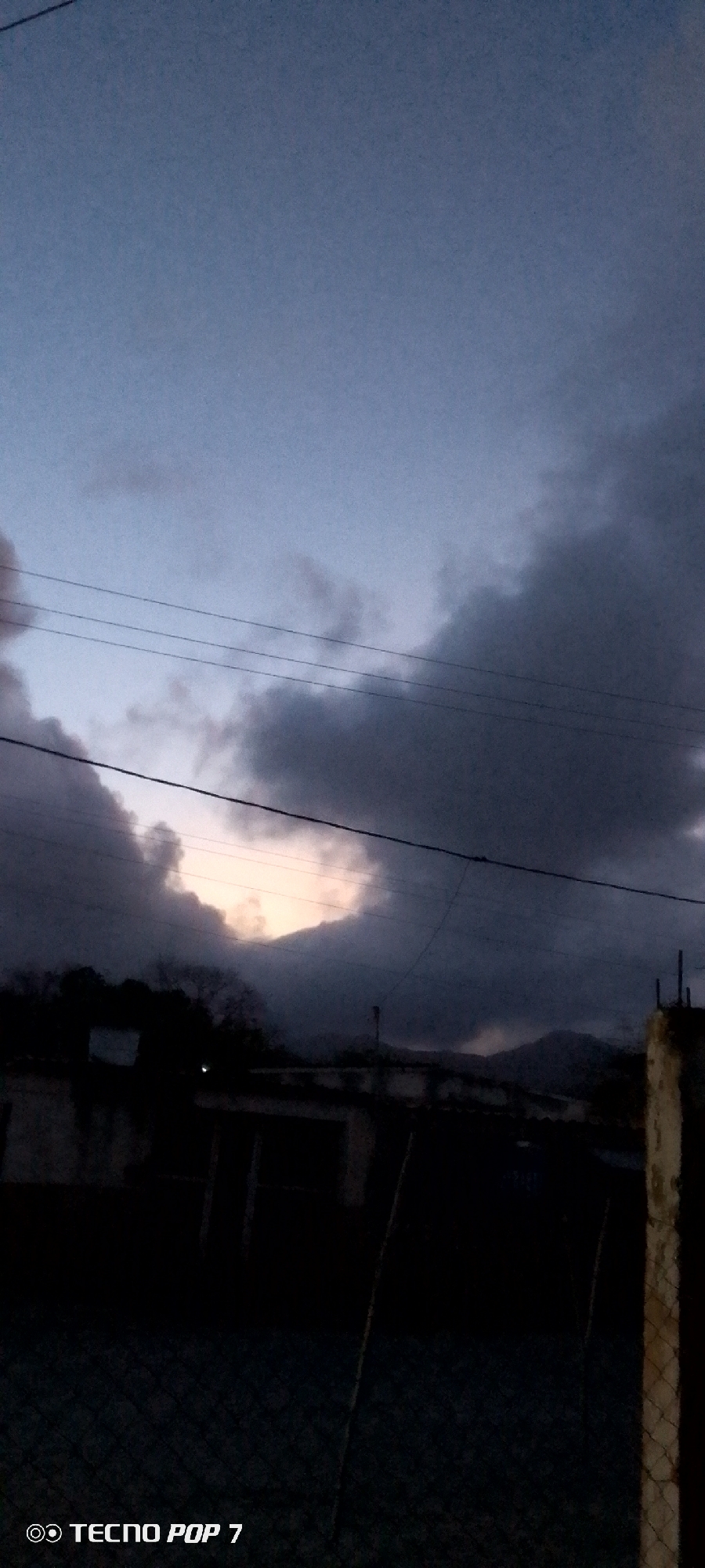 The image depicts a cloudy, dark sky over a building in the city of Altagracia, Venezuela. There are electrical wires visible in the sky, and a mountain or hill can be seen through a break in the clouds. A chain-link fence is in the foreground. The image was taken with a TECNO POP 7 smartphone.
