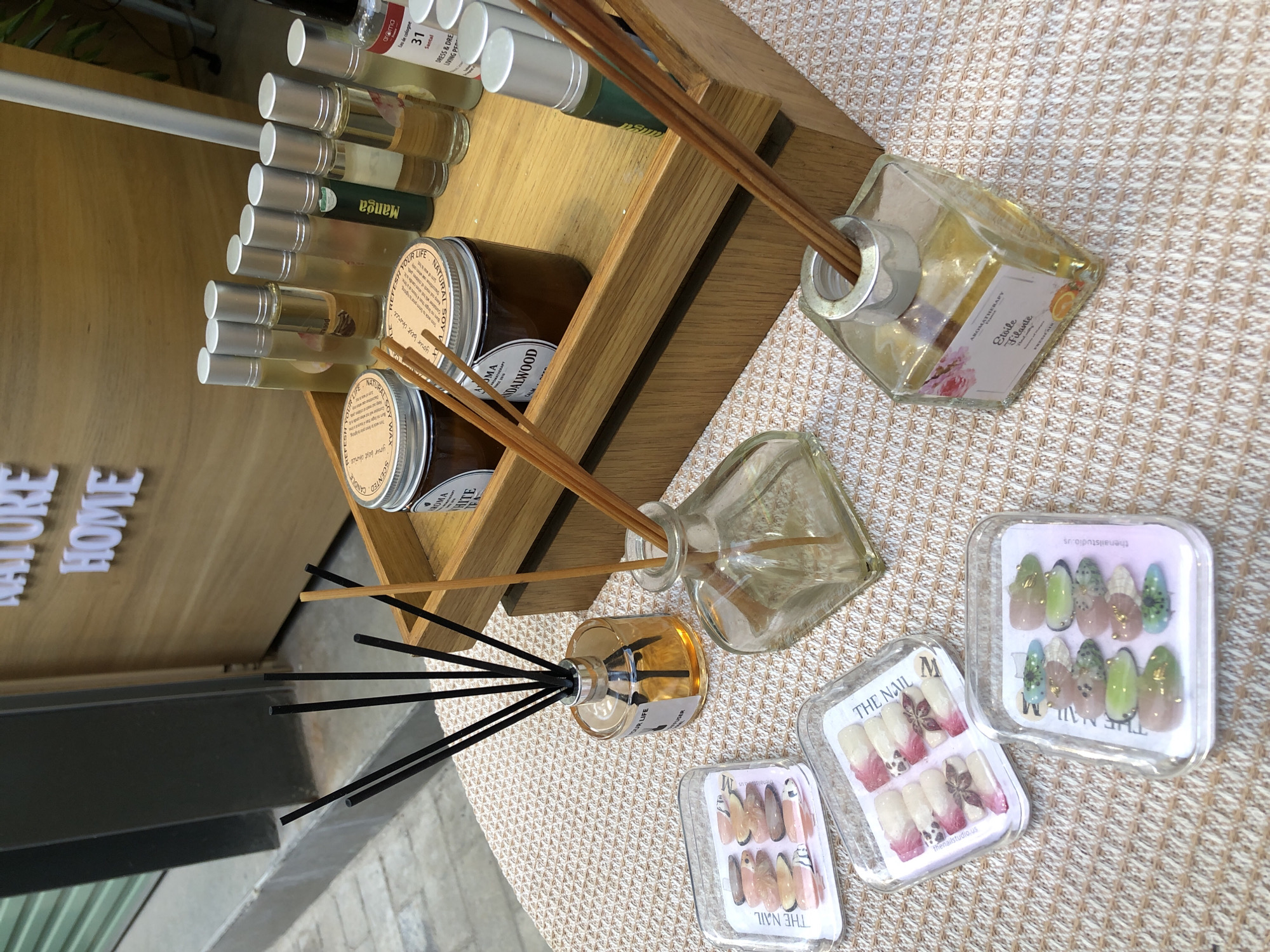 The image depicts a display of scented products and artificial nails at an outdoor market stall. In the background, a wooden sign reads "NATURE HOME" in white lettering. The foreground features a textured peach-colored tablecloth.
Several glass diffusers are visible, some filled with amber-colored liquid and reeds. One diffuser, positioned on the left, has a label that is partially obscured but includes the words "YOUR LIFE." Another diffuser on the right has a label reading "AROMATHERAPY," "Etoile," "Filante," and "FRESH AIR." Two glass jars with lids are also present; one has a label with text that includes "SANDALWOOD."
A collection of small, clear plastic containers holds artificial nails. These containers are arranged in a row on the tablecloth. The nails display various designs, including floral patterns, ombré effects, and what appear to be small charms or embellishments. One container has the branding "THE NAIL" printed on it.
A wooden tray holds several small glass bottles with silver caps. Some of these bottles are filled with a pale yellow liquid. One bottle has a label with the word "Manga" in green lettering. Another bottle has a label with "31 SANTAL" and "DRESS & DRE..." visible.
The overall impression is a retail display of beauty and home fragrance products, likely in a market or shop setting. The backdrop suggests an urban environment, possibly in Đà Nẵng, Vietnam, given the context of the prompt. There are no people visible in the image.