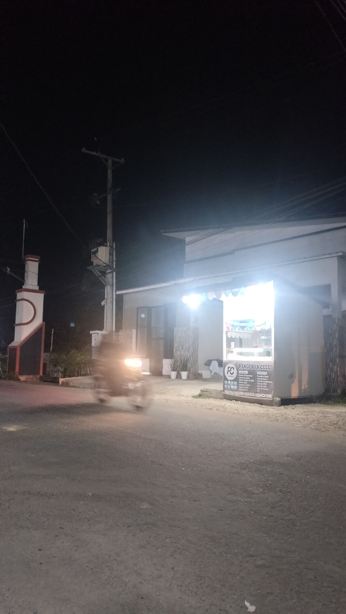 The image depicts a street scene at night in Lhokseumawe, Indonesia, likely in a commercial or residential area. A brightly lit storefront, identified by a sign as "PANGERAN CELL," is visible on the right side of the frame. The storefront appears to be a small kiosk or shop selling mobile phone services and possibly accessories, indicated by a poster detailing various mobile data packages and services. To the left of the storefront, a monument or marker with a distinctive white, red, and black design stands prominently. A motorcycle is captured in motion, blurred due to the long exposure, with its headlights illuminated, moving on the asphalt road. Power lines and a utility pole are visible in the background, common in urban Indonesian landscapes. The overall atmosphere is nocturnal, with artificial lighting from the storefront and vehicle headlights illuminating the otherwise dark surroundings.