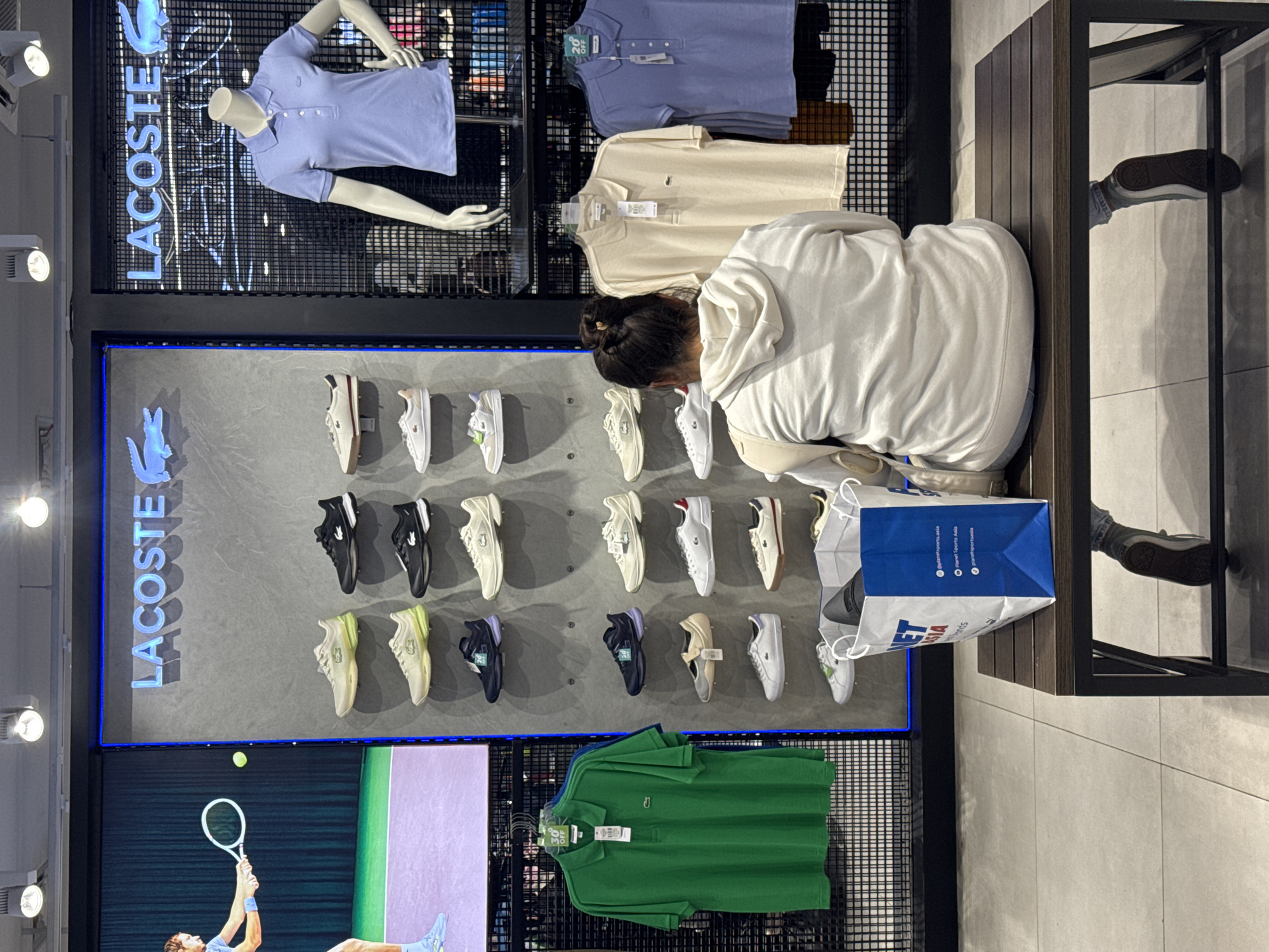 The image depicts the interior of a Lacoste store, likely located in Kuta, Indonesia, given the presence of a "Planet Sports Asia" shopping bag. In the foreground, a person wearing a white hoodie and jeans is seated on a wooden bench, facing away from the camera, looking towards a display of Lacoste clothing and shoes. The person is holding a white and blue shopping bag on their lap.
The background features a well-lit display area. On the left, a large screen shows a tennis player in action. Adjacent to this screen is a wall with a large "LACOSTE" logo and several shelves displaying rows of Lacoste sneakers in various colors, including white, beige, and black. Further to the right, another "LACOSTE" sign is visible, with a mannequin dressed in a light purple Lacoste polo shirt. Below the mannequin, more Lacoste polo shirts are displayed on racks in beige and blue. The floor is tiled in a light grey pattern. The overall environment suggests a retail setting focused on athletic and casual wear.