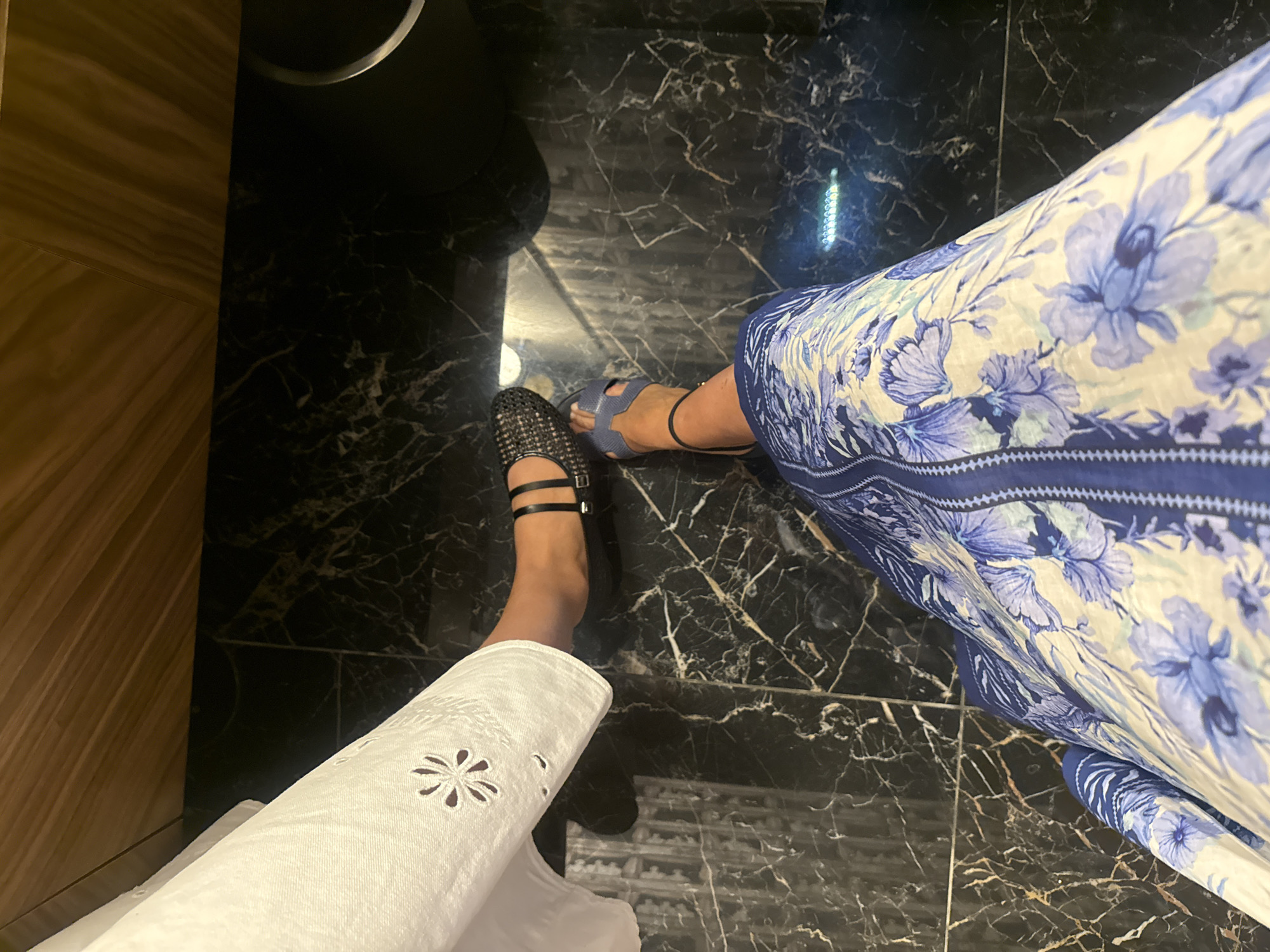 The image shows a view looking down at two pairs of feet standing on a dark marble floor. To the left, a foot is wearing a black sandal with multiple straps. The person is wearing a white garment with decorative cutouts. To the right, a foot is wearing a blue sandal. The person is wearing a long dress with a floral pattern in blue and white. The background includes a wooden surface on the left and a black object near the top. The location is unknown but stated to be Vila Verde, Portugal.