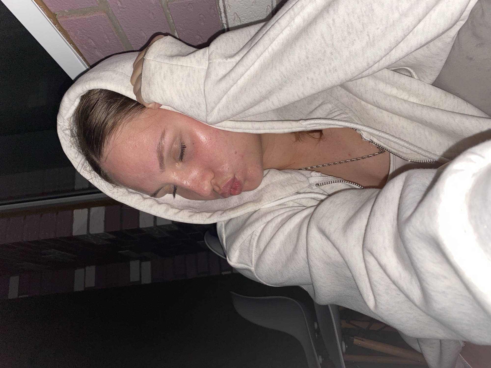 The image shows a close-up of a young person wearing a light-colored hooded sweatshirt and a silver chain. The person's eyes are closed, and their lips are pursed as if they are about to kiss. Their face appears flushed, and there are visible pores and some redness on their cheeks. The person is leaning their head on their hands, which are inside the hood of the sweatshirt. The background is dark, with a hint of a brick wall and a window visible on the right side. There is also a blurry image of a chair in the lower left corner. The scene appears to be outdoors at night. The metadata indicates the image was taken in Izhevsk, Russia.