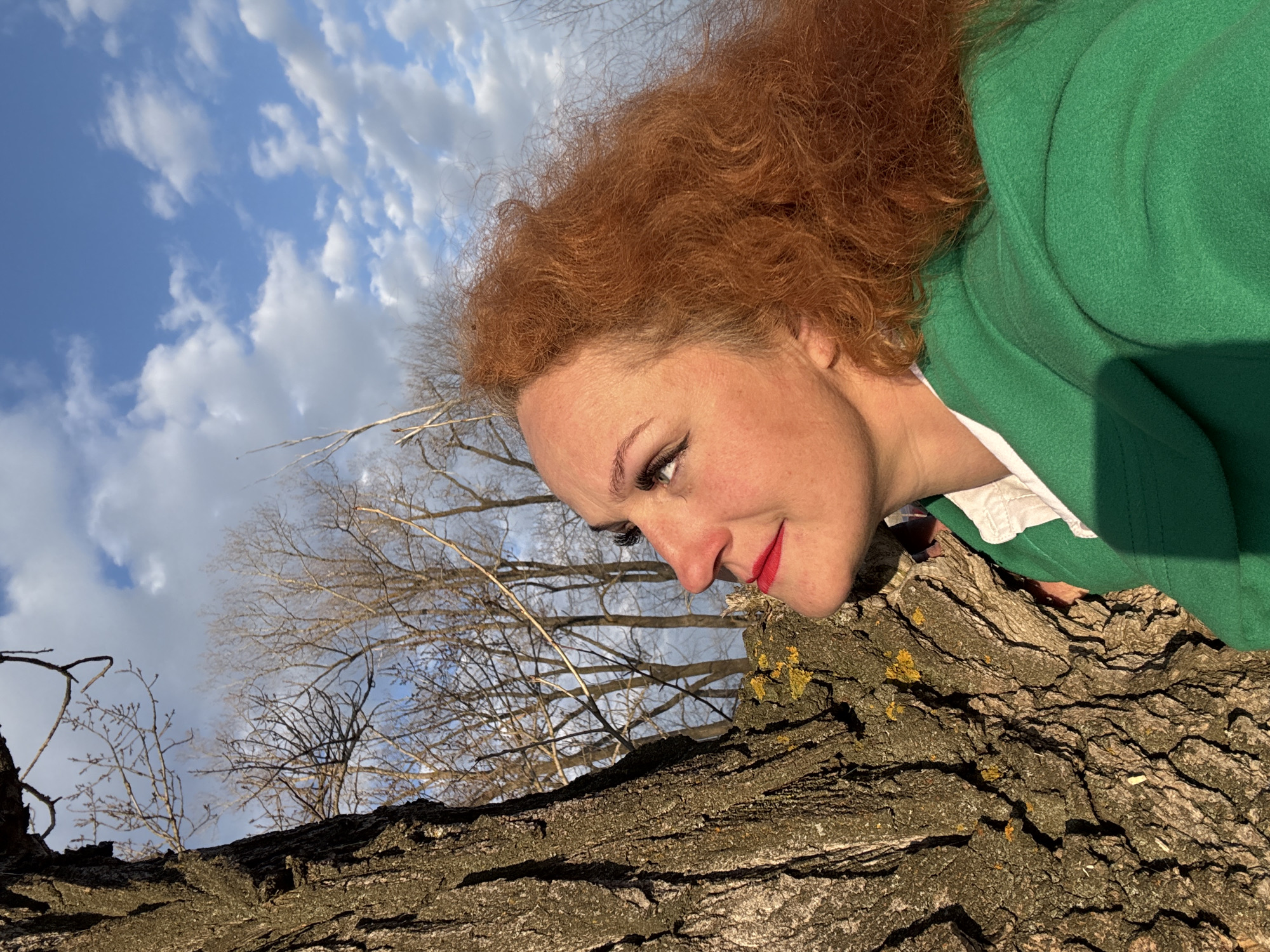 A woman with red, curly hair, wearing a green jacket over a white shirt, is positioned to the right of the frame, with her face turned downwards and slightly to the left. She has red lipstick and appears to be resting her face near a large tree trunk that runs horizontally across the lower portion of the image.
Behind the woman and the tree trunk, there's a view of bare tree branches extending upwards towards a blue sky filled with scattered white clouds. The light suggests it is daytime, possibly late afternoon. The tree trunk shows visible bark texture and some signs of lichen growth.
It is not possible to determine the location as the city of Noviy, Russia. The background does not provide sufficient information.