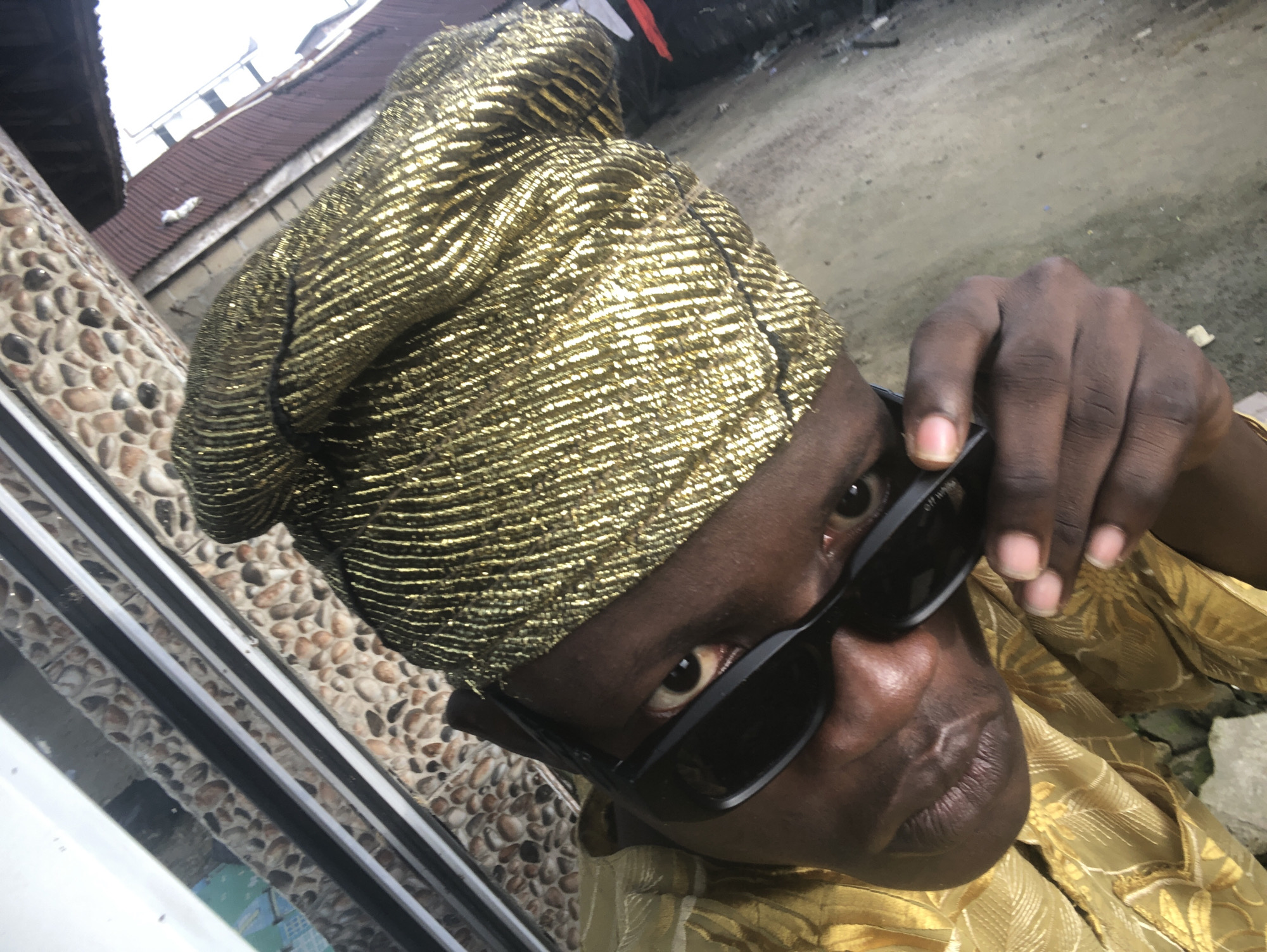 The image shows a person wearing a golden head wrap and a golden outfit. The person is wearing black sunglasses and is holding them with their right hand. The background appears to be an outdoor setting, with a wall made of stones and a corrugated iron roof visible. The ground is dirt or gravel. The scene is tilted, suggesting the photo was taken at an angle. The overall impression is of a close-up portrait with a candid feel, set against a somewhat urban or semi-urban backdrop in Ado, Nigeria.