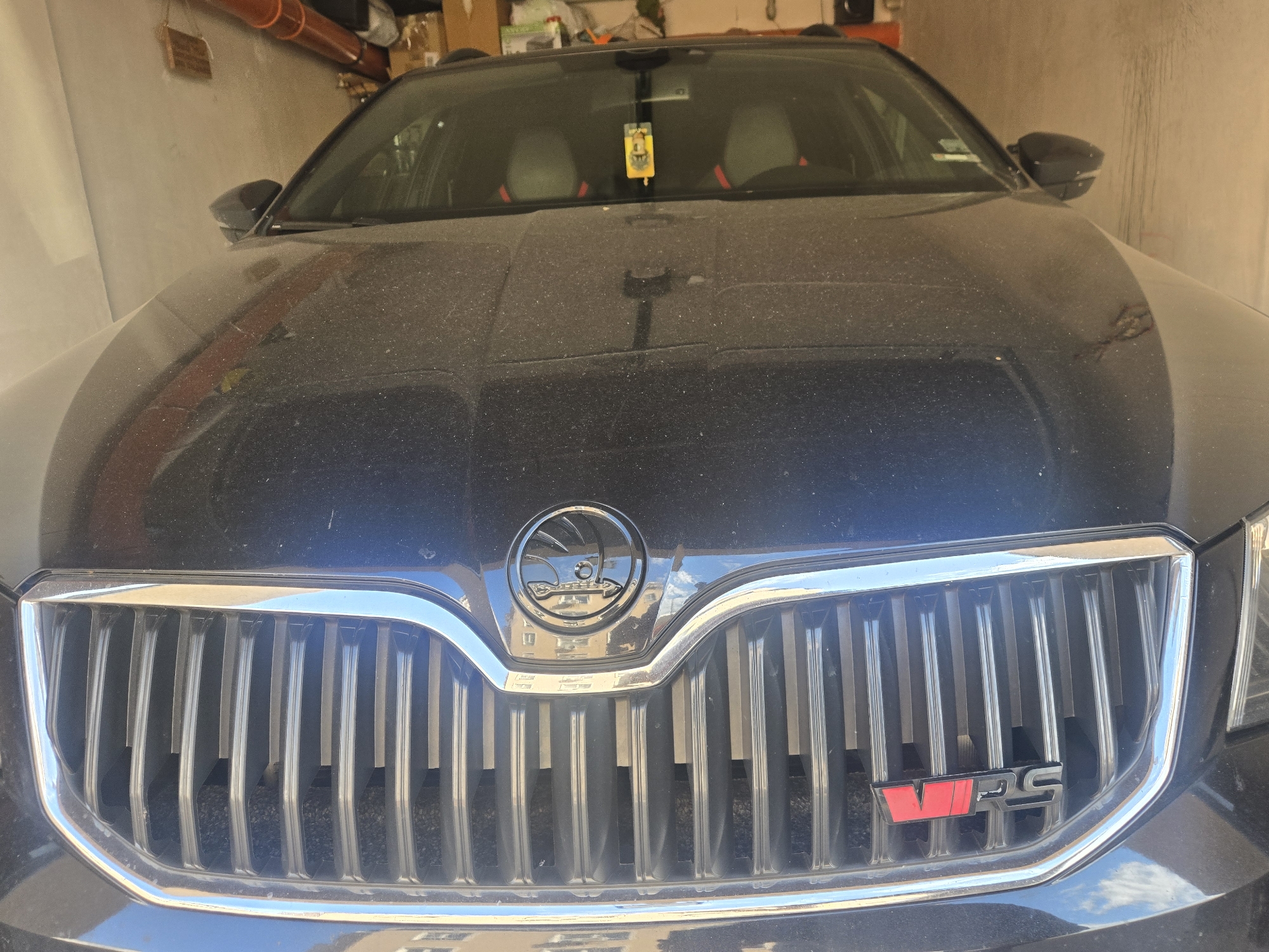 The image shows a close-up of the front of a dark blue Skoda Octavia vRS car. The car appears to be parked indoors, possibly in a garage, as suggested by the wall behind it. The car is dusty. The focus is on the front grille, which features the Skoda logo and the "vRS" badge. The interior of the car is visible through the windshield, showing the seats and dashboard. There is a small air freshener hanging from the rearview mirror. The location appears to be Unknown, Unknown.