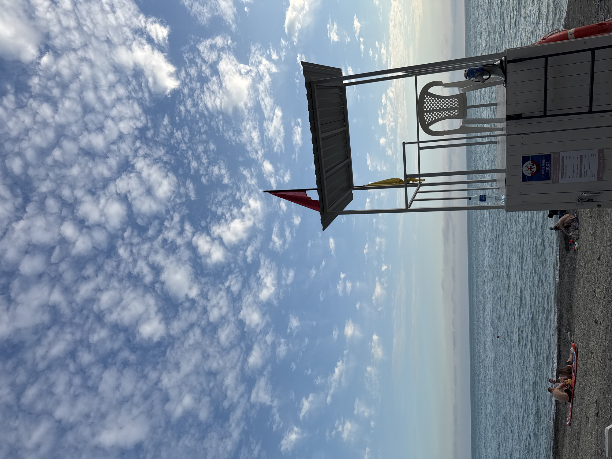 A lifeguard tower stands on a pebble beach next to a calm, blue sea under a sky filled with scattered white clouds. A red flag flies from the top of the tower, and a yellow flag is visible inside. A white plastic chair is placed on the platform, along with a water bottle and a megaphone. The tower has a white base with a ladder leading up to the platform. A sign on the base displays text and a logo. On the beach, a few people are seen relaxing on a red inflatable mat in the foreground, while others are further down the beach. In the distance, the horizon is clearly defined where the sea meets the sky. The overall atmosphere is peaceful, suggesting a typical day at the beach. The specific location within Sochi, Russia, cannot be determined from the image alone.