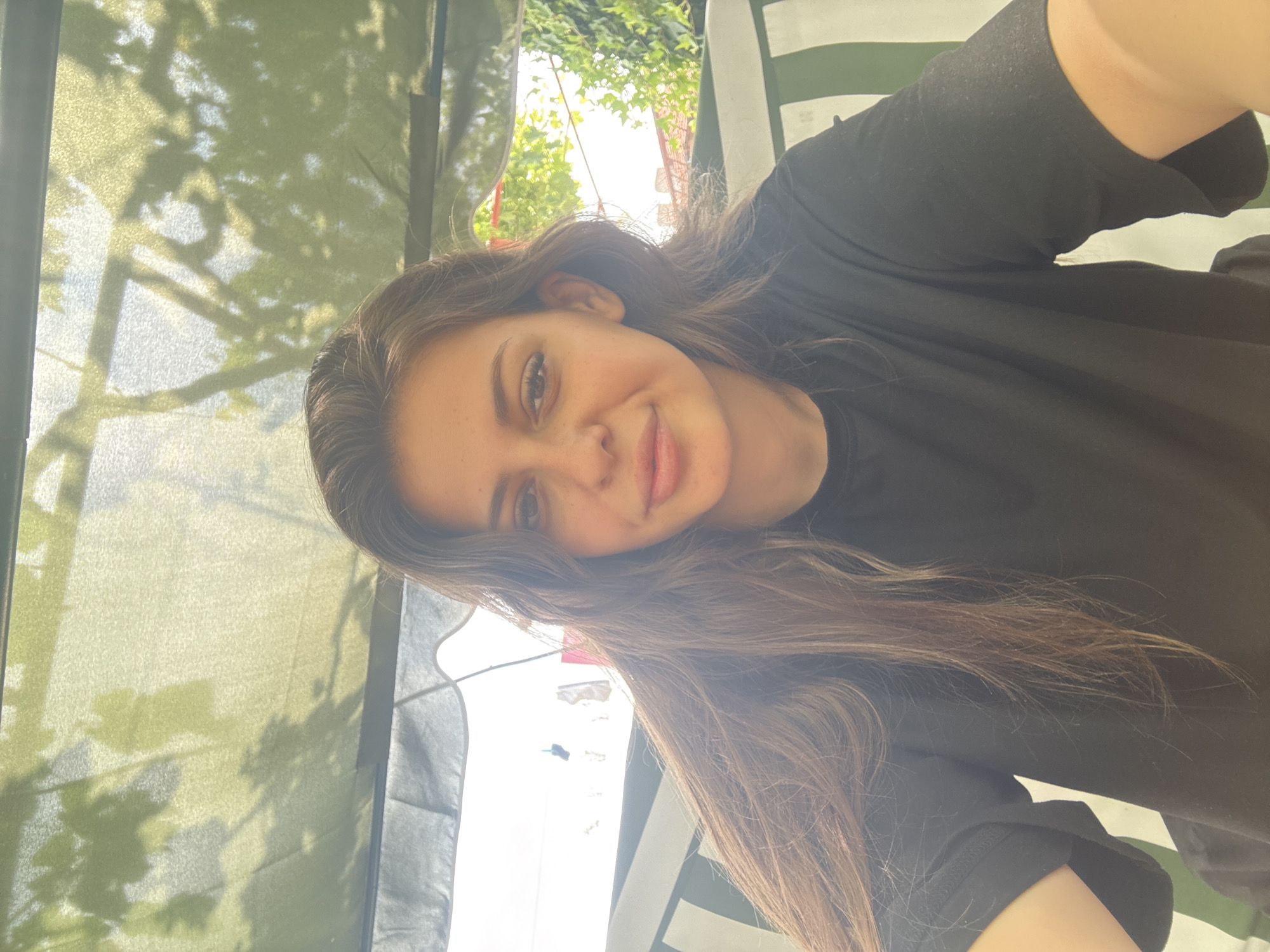 A young woman is taking a selfie outdoors, presumably in Govedare, Bulgaria, as indicated by the context of the request. She is wearing a black t-shirt and has long, brown hair. The background suggests an outdoor setting, possibly a garden or patio, with some greenery visible and sunlight filtering through. There appears to be some fabric overhead, possibly a canopy or awning, with shadows of leaves cast upon it. The overall lighting is bright, suggesting it is daytime. The woman's expression is relaxed and she is looking directly at the camera.