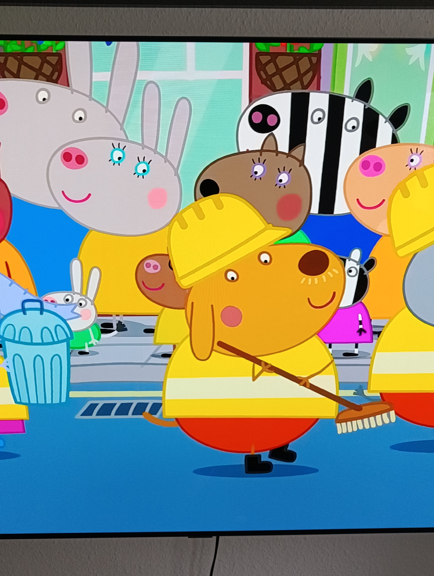 The image depicts a scene from the animated children's show "Peppa Pig". It shows an array of cartoon characters including Peppa Pig herself, a donkey, a zebra, and a dog. The dog, in the foreground, is wearing a yellow hard hat and a striped yellow and white shirt. He is also wearing red pants and black shoes and is holding a broom as if sweeping. Beside him is a garbage can and what appears to be the bottom of a fire hydrant. The scene is set on a city street with the characters seemingly part of a sanitation crew.