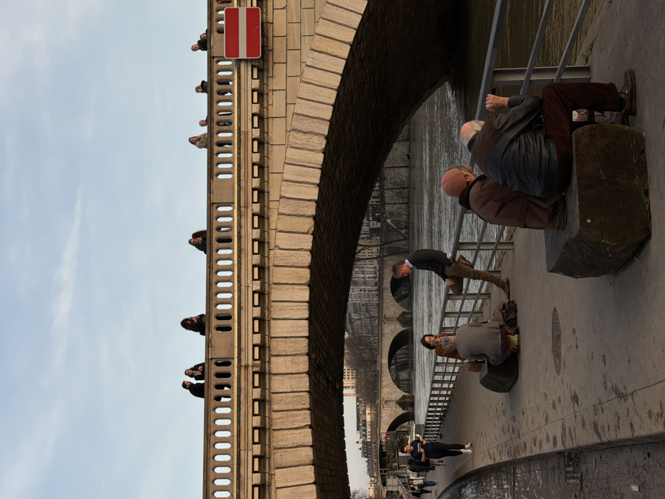 The scene depicts a clear, late afternoon or early evening view along the Seine river in Paris, France, specifically beneath a large stone arch bridge.
In the foreground, a paved quayside runs parallel to the river, bordered by a metal railing. On the lower right, two older individuals are seated on a rectangular stone bench, facing the river with their backs mostly to the viewer; one wears a brown jacket, the other a dark leather jacket. To their left, a man in a dark sweater and brown trousers leans against the metal railing, facing a group of three. This group includes a woman with dark hair in a brown jacket seated on another stone bench, and another person in a light grey jacket seated on the ground nearby. Further left on the quayside, a younger individual in a dark shirt and light pants walks away from the viewer, looking at a mobile phone. Additional indistinct figures are walking in the distance along the quayside.
The dominant feature in the midground is a large stone arch bridge spanning the river. Its upper structure features an ornate balustrade with repeating circular openings. Multiple individuals are visible above this balustrade, silhouetted against the light blue sky, some appearing to observe the surroundings, others walking. One person in the center of the bridge appears to be looking at a phone. A red and white circular "No Entry" (Sens Interdit) traffic sign is prominently affixed to the right side of the bridge's stone structure.
In the background, two more stone arch bridges are visible further down the river. The distant riverbanks are lined with multi-story buildings characteristic of Parisian architecture, featuring light-colored stone facades. The sky is clear blue with faint cloud formations. The river water is calm and appears brownish.