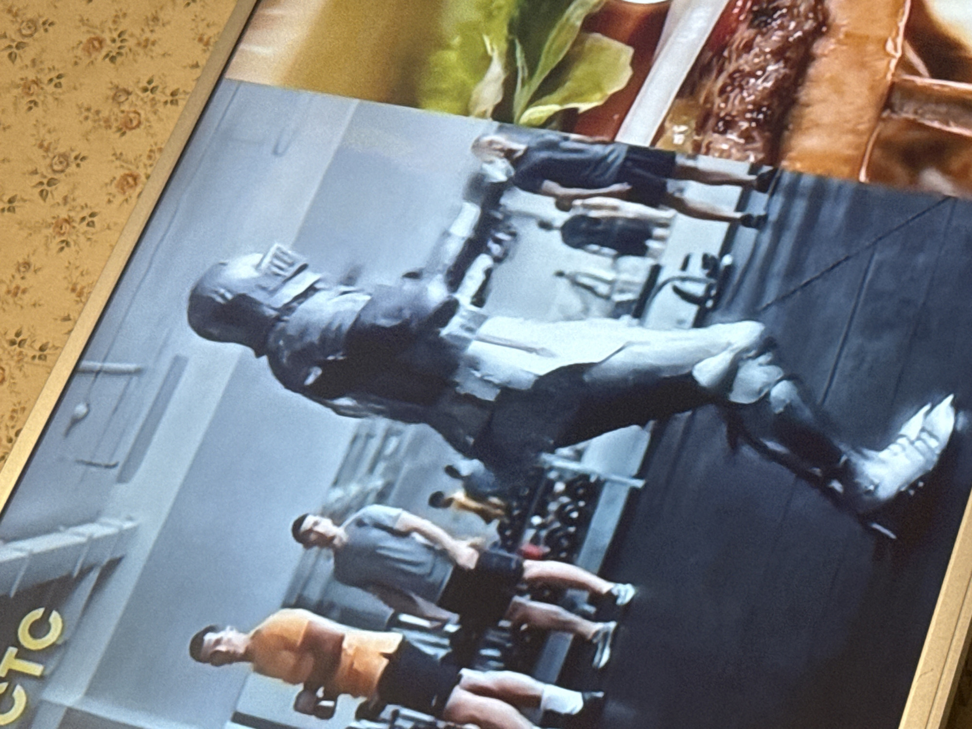 The image depicts a poster featuring a digitally rendered character, possibly from a video game, superimposed over a gym scene. In the foreground, there are several men performing weightlifting exercises. The gym setting includes weight racks and a minimalist aesthetic. A partial logo is visible on the bottom left corner, displaying the letters "TC". The location of the gym in Mytishchi, Russia, is not determinable from the image.