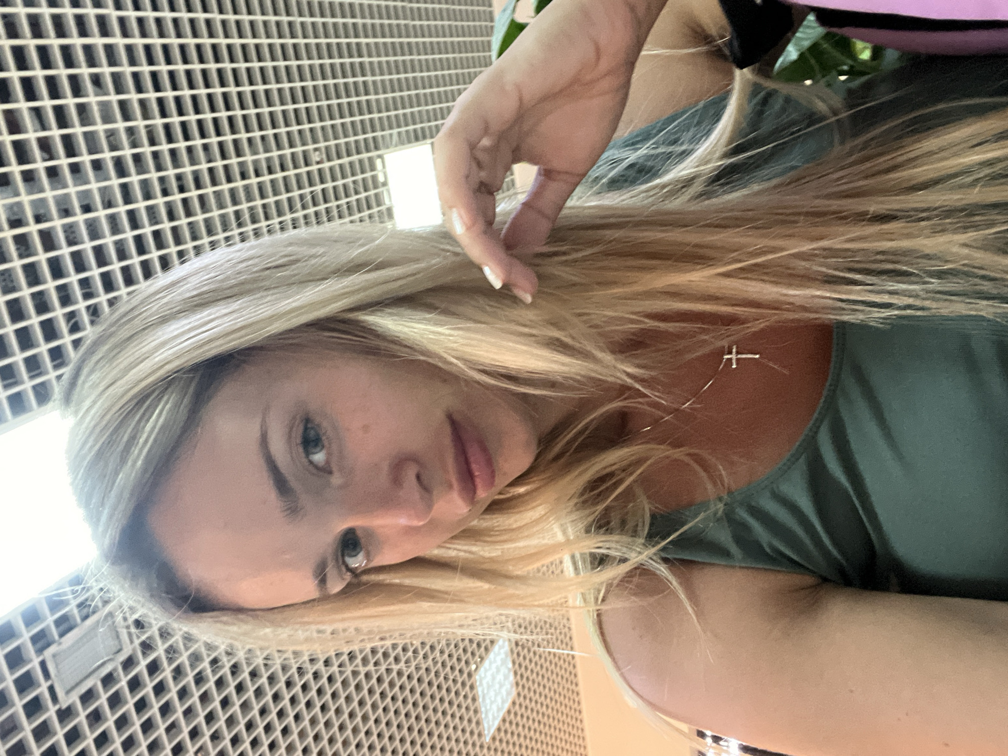 The image shows a close-up, selfie-style portrait of a woman with blonde hair, wearing a green tank top and a silver cross necklace. She is indoors, with a grid-patterned ceiling and fluorescent lighting visible in the background. Her right hand is near her face, touching her hair. The lighting suggests an indoor public space, potentially an airport or a shopping mall, but there are no specific visual cues to pinpoint a location within Chișinău, Moldova. The woman's expression is neutral, and she is looking towards the camera.