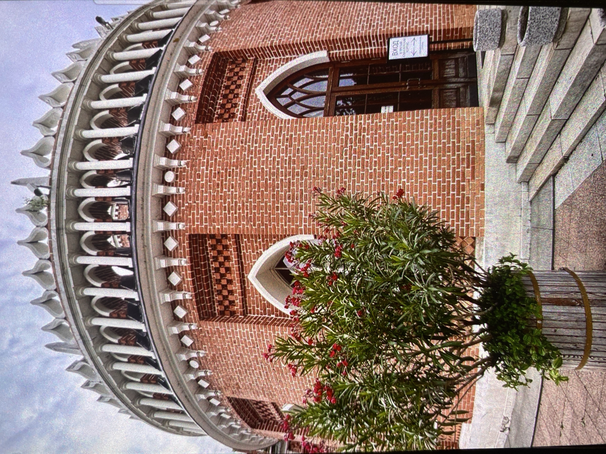 The image shows a section of a circular brick building, likely located in Saint Petersburg, Russia, given its architectural style. The building features two arched windows with wooden frames, brickwork in varying patterns and colors, and a white decorative colonnade around the top. A sign with Cyrillic text hangs above one of the windows, possibly indicating the entrance. A potted plant with green leaves and red flowers sits near the base of the building. Adjacent to the building is a gray stone structure with steps. The perspective is slightly tilted, with the sky visible in the upper left.