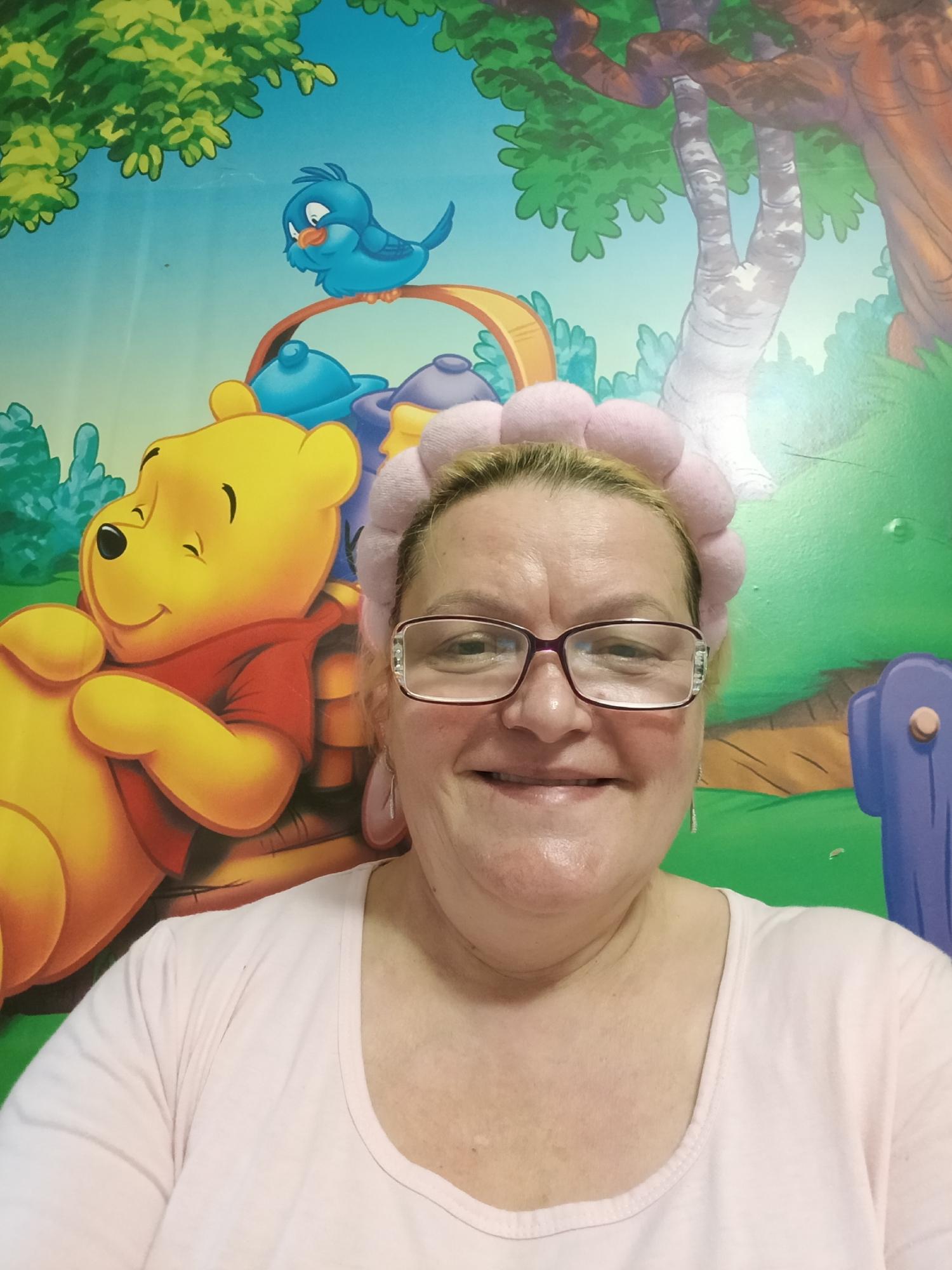 A woman is centered in the frame, wearing a pink headband and glasses, against a backdrop of a Winnie the Pooh mural. The mural includes Winnie the Pooh, a blue bird, trees, and other cartoon elements. The woman wears a light pink top. The location is possibly Красный Маяк, Russia, but this detail is unconfirmed from the image alone.
