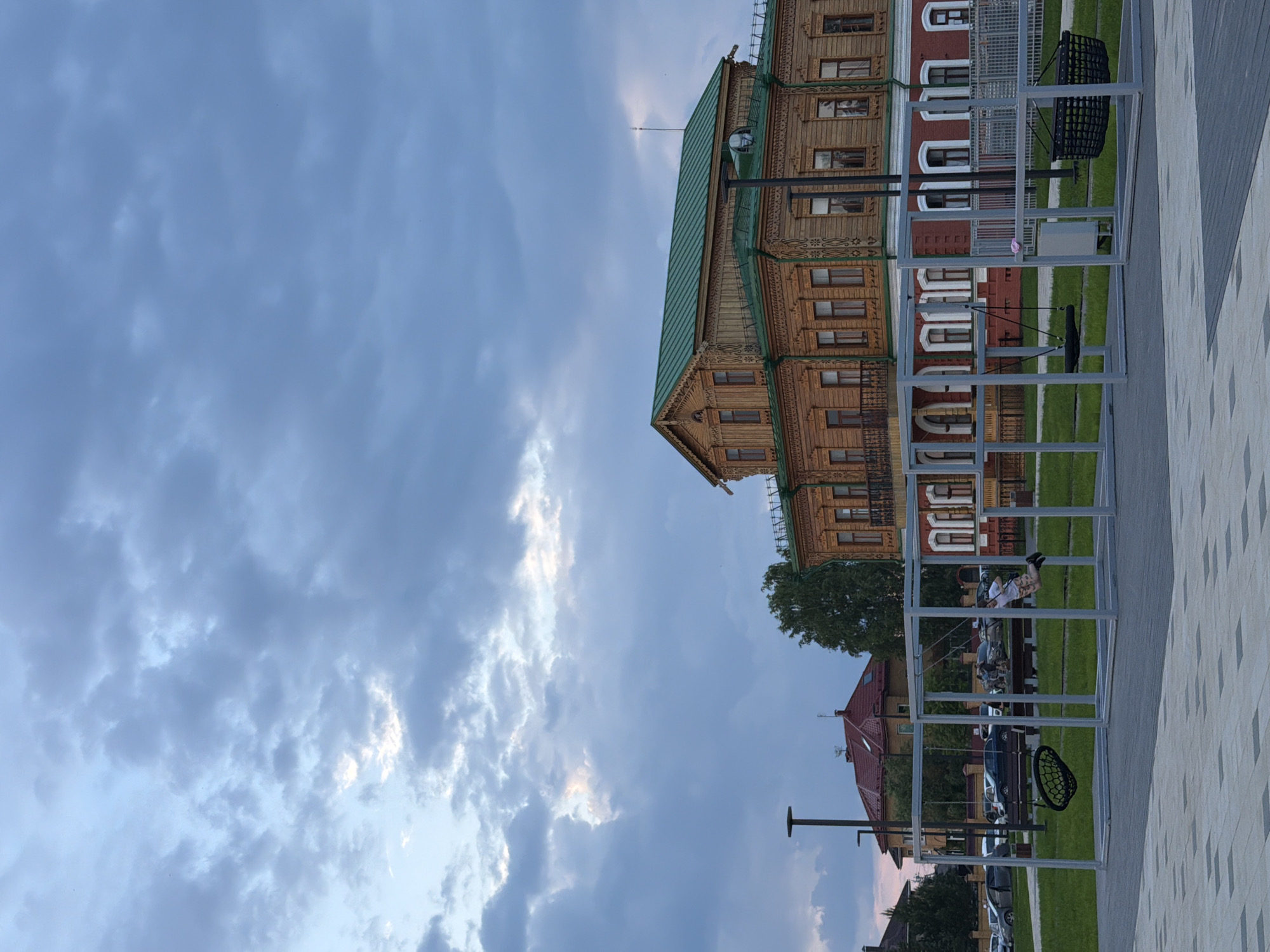 The image depicts a public park area in Solikamsk, Russia, featuring a swing set in the foreground and a large, intricately decorated wooden building in the background. The sky above is filled with dramatic, cloudy formations in shades of grey and white, suggesting dusk or an overcast day.
In the middle ground, a person is actively using a swing, their body arced backward in motion. The swing set is a modern, metallic structure with two distinct swings: one a traditional seat swing and the other a basket-style swing. Adjacent to the swing set, a paved area with a patterned surface is visible.
The building behind the park area is a prominent two-story structure constructed primarily of wood. It is characterized by detailed ornamental carvings along its facade, particularly around the windows and roofline. The building has a green, sloped roof and numerous windows with dark frames. In front of this wooden structure, a portion of a red-brick building with white-trimmed windows is also visible.
Beyond the park and buildings, several parked cars are discernible, along with some trees and lampposts, indicating an urban or semi-urban setting. The overall impression is a serene public space with a blend of historical architecture and modern recreational facilities.