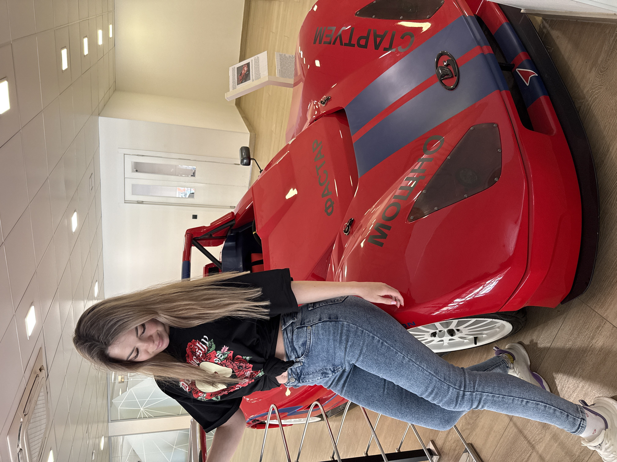 A person with long blonde hair is leaning against a red race car in what appears to be an indoor exhibit space. The person is wearing a black t-shirt with a floral design, blue jeans, and white sneakers with purple accents. The red race car has blue stripes and text in a foreign language on its body. The exhibit space has a clean, modern design with white walls and ceiling, and appears to be in the city of Unknown, Unknown. There's also a display with printed material on a stand behind the car. The floor is a light wood-grain pattern.