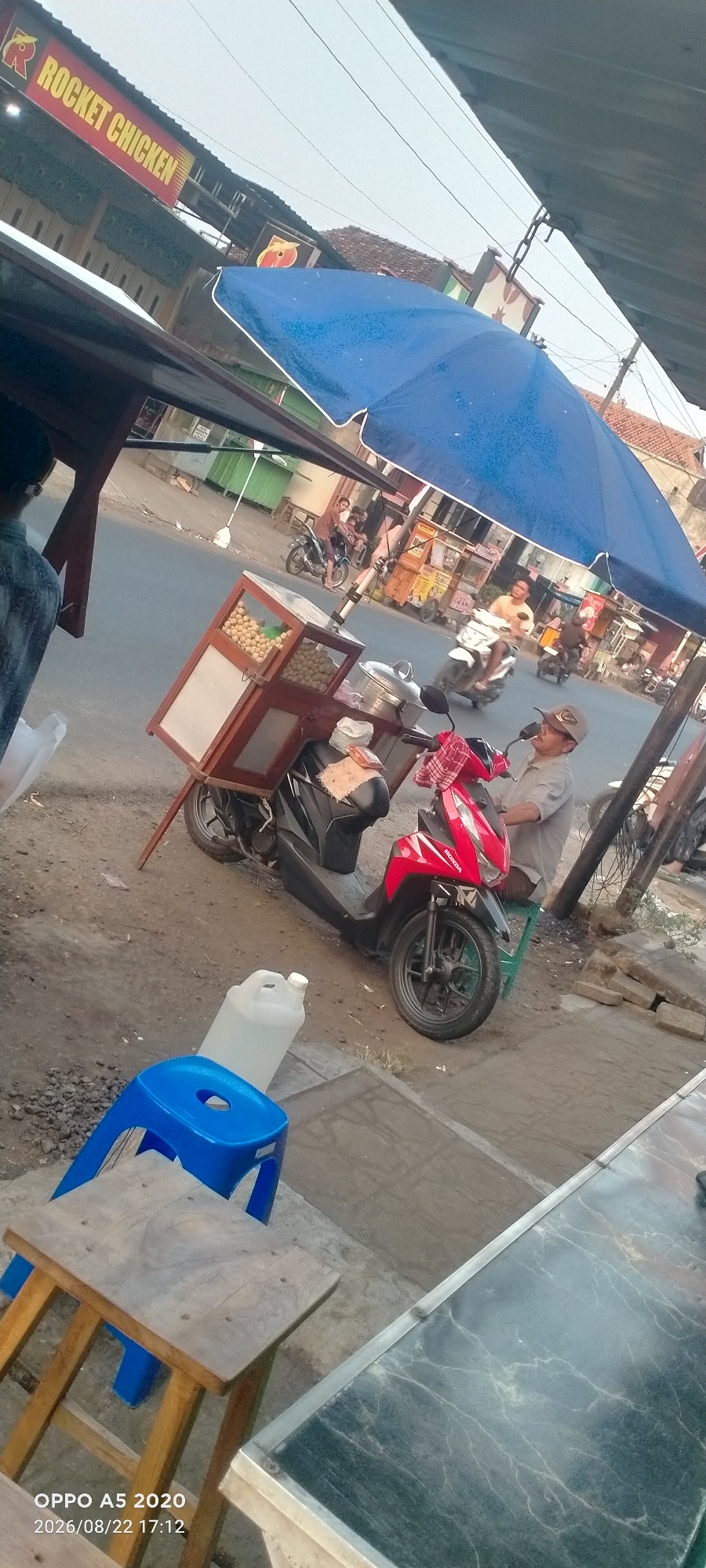 The image depicts a street scene in Kedung, Indonesia, featuring a mobile food cart. The food cart is attached to a red and black scooter. It has a wooden frame with a glass display case filled with round food items, possibly snacks or sweets. A metal pot with a lid is also visible on the cart. A man wearing a cap and a grey shirt is seated on a green stool behind the scooter, seemingly operating the cart. In the foreground, a blue plastic chair and a wooden stool are placed on the ground. A large white plastic water jug is positioned behind the blue chair. Further in the background, other people on motorcycles and food stalls can be seen along the street, with buildings and a blue umbrella suggesting a marketplace or busy road. A prominent red sign with yellow lettering that reads "ROCKET CHICKEN" is visible on a building in the upper left corner of the image. The overall setting appears to be an outdoor market or roadside vendor area.