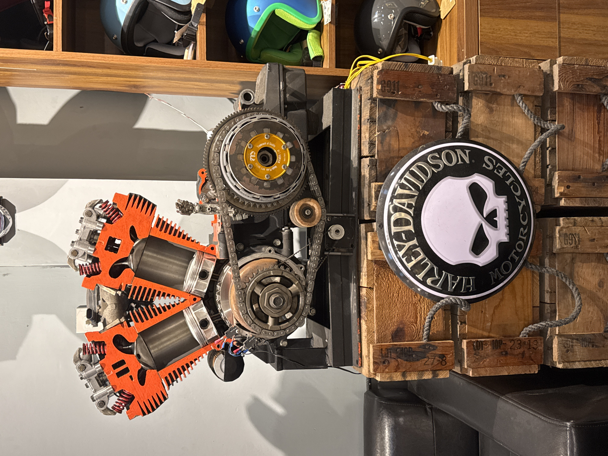 The image depicts a detailed cutaway display of a V-twin motorcycle engine, prominently featuring its orange cylinder heads and silver pistons. The engine is mounted on a black metal stand atop a stack of weathered wooden crates. A circular Harley-Davidson logo with a skull emblem is attached to the front of the crates. In the background, a wooden shelving unit displays several motorcycle helmets of various colors, including blue and gray. To the left, a dark leather armchair is partially visible. The overall setting appears to be an indoor retail or display space, possibly a motorcycle dealership or a themed cafe, located in Nha Trang, Vietnam, as suggested by the context. There are no people visible in the image. The lighting suggests an indoor environment, possibly with artificial lighting contributing to the scene's illumination.