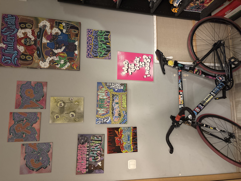 A gray wall is adorned with nine vibrant art pieces, predominantly graffiti-style paintings, alongside a bicycle leaning against the wall. The paintings vary in color and content, ranging from abstract letterings to cartoon-like characters. The bike is mostly black with colorful stickers decorating the frame and red rims. A light switch is noticeable on the lower right part of the wall. A shelf with various items on top can be seen above the paintings.