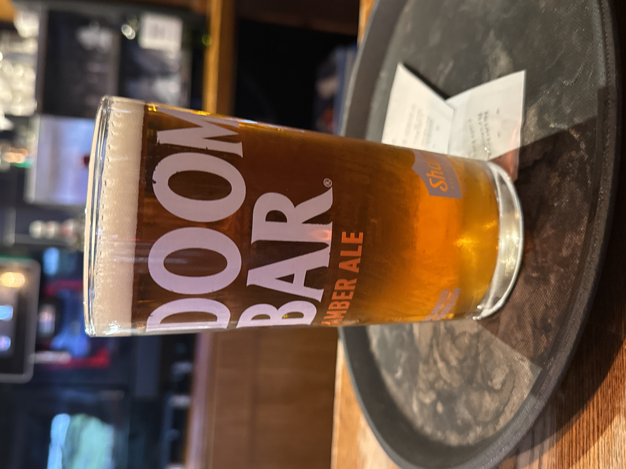 The image shows a pint glass of Doom Bar Amber Ale on a black serving tray. A receipt sits nearby on the tray. The background is blurred, suggesting a bar or restaurant setting. The location is Great Yarmouth, United Kingdom, based on the orange logo on the glass.