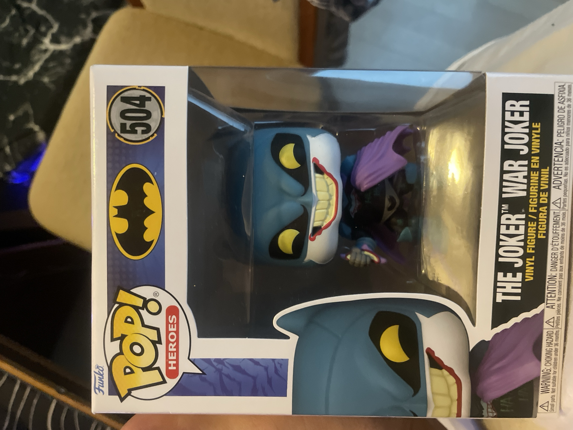 A close-up photograph depicts a Funko Pop! Heroes figurine of "The Joker War Joker" in its packaging. The box features the Funko Pop! logo with the word "Heroes" underneath, a bat symbol, and the number "504" in a circle. The figurine inside is a stylized representation of The Joker. Text on the box includes the character's name and warnings in English, French, and Spanish. The box is positioned on a surface in an indoor setting. There is no indication of Lachendorf, Germany or its surroundings.