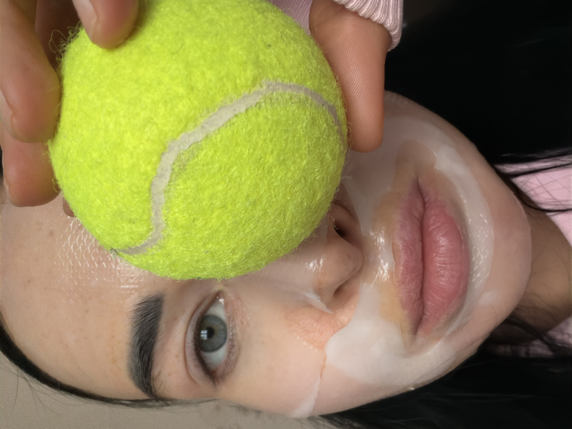 A close-up, rotated image depicts a person with a white face mask, covering most of their face except for the eye, eyebrow, and lips. A bright yellow tennis ball, held by a hand, is positioned over the person's forehead. The person appears to be wearing a light pink garment. The background is dark. The location is not discernible from the image.