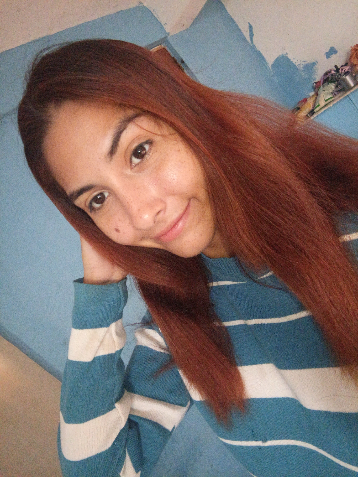 The image shows a woman with reddish-brown hair resting her head on her hand. She has freckles and is wearing a blue and white striped sweater. The background is a light blue wall. There is no information in the image to support the location being in Juan Griego, Venezuela.