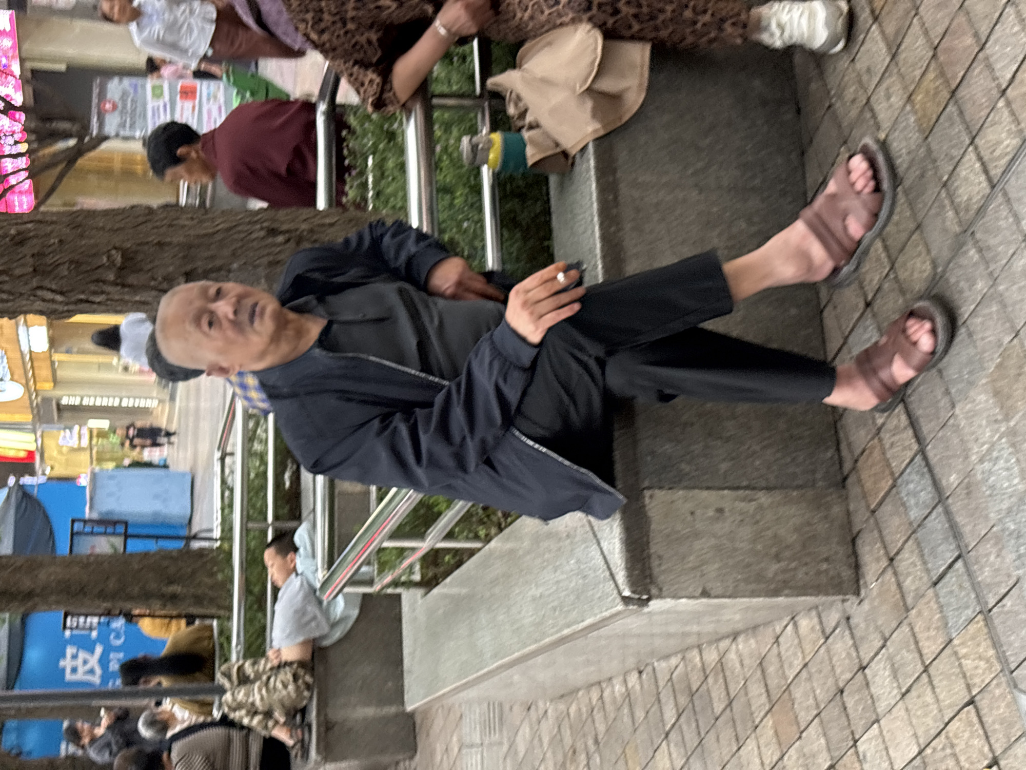 This photograph depicts a street scene in Jiangjin, China, focusing on several individuals. In the foreground, an elderly man with a bald head is seated on a concrete bench, wearing a dark jacket over a grey t-shirt and black trousers. He is wearing brown sandals and appears to be holding a cigarette. To his right, a woman in a leopard-print dress and white sneakers is partially visible. To the left of the elderly man, another individual, possibly a younger person, is lying down on the same bench, wearing camouflage-patterned pants and a light grey t-shirt, with their head resting on their arm. In the background, other people are seen walking and standing. The environment includes trees, railings, and a tiled pedestrian walkway. A blue sign with Chinese characters is visible behind the person lying down, and a pink neon sign is also apparent in the distance. The overall setting suggests a public urban space.