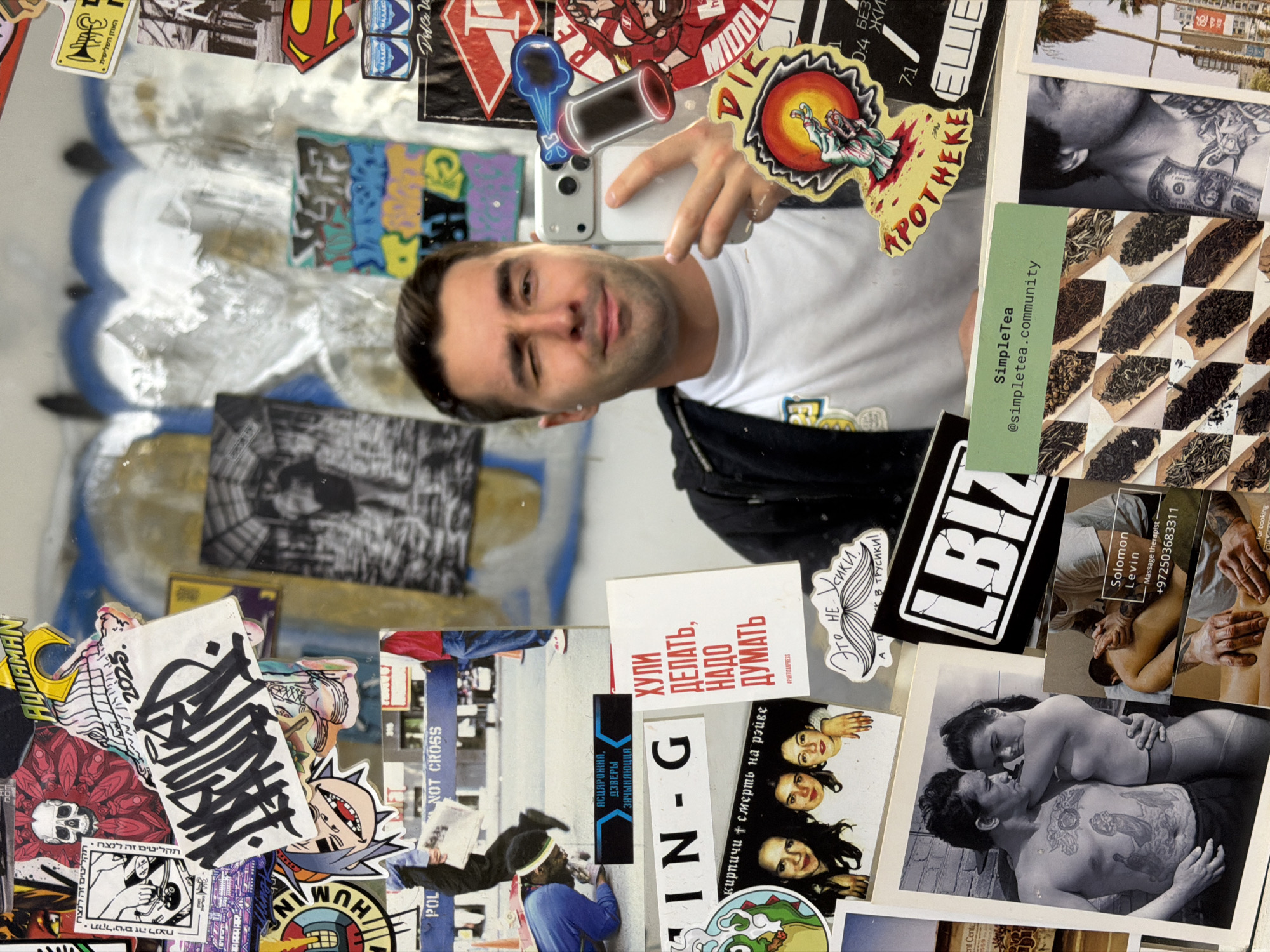 A male in his late 20s to early 30s is depicted taking a selfie in a reflective surface, winking with his right eye, and holding a light-colored smartphone with his right hand. He wears a white t-shirt and a dark jacket. The background is a dense collage of numerous stickers, photographs, and graffiti on a wall, strongly suggesting a public or commercial establishment.
Key details within the collage include:
* **Graffiti and Stickers:** Patches of blue and white spray paint are visible. Prominent stickers include a skull, an "AQUAMAN" logo, various stylized graffiti tags (one with "2025"), a Gorillaz character, and a Superman logo. Russian text appears on a sticker reading "АСЦАРОЖНА, ДЗВЕРЫ ЗАЧЫНЯЮЦЦА" (Caution, doors closing) and on a white card with red text stating "ХУЛИ ДЕЛАТЬ, НАДО ДУМАТЬ" (What to do, need to think), alongside "#PARTISANPRESS". Another sticker features a mustache and Russian text "ЭТО НЕ УСИКИ, А УЛЫБКА В ТРУСИКИ!" (These are not mustaches, but a smile in panties!). A black sticker with "LBIZ" in white and a yellow "APOTHEKE" sticker with a stylized hand are present.
* **Photographs:** Several photos are integrated into the collage: a street scene with a "POLICE DO NOT CROSS" barrier and a person kneeling, a group of three women, a black and white image of a shirtless man with tattoos embracing a woman, and a cropped black and white image of another tattooed, shirtless man.
* **Business Cards:** A green card for "SimpleTea @simpletea.community" displays various types of dried tea leaves. A business card for "Solomon Levin Massage therapist" with the Israeli phone number "+972503683311" and images of massage is also affixed.
The presence of an Israeli phone number prefix (+972) and significant Russian language elements strongly indicates the location is in Israel, consistent with the diverse urban environment of Tel-Aviv. The overall aesthetic suggests an artistic, urban, or counter-culture space within the city.