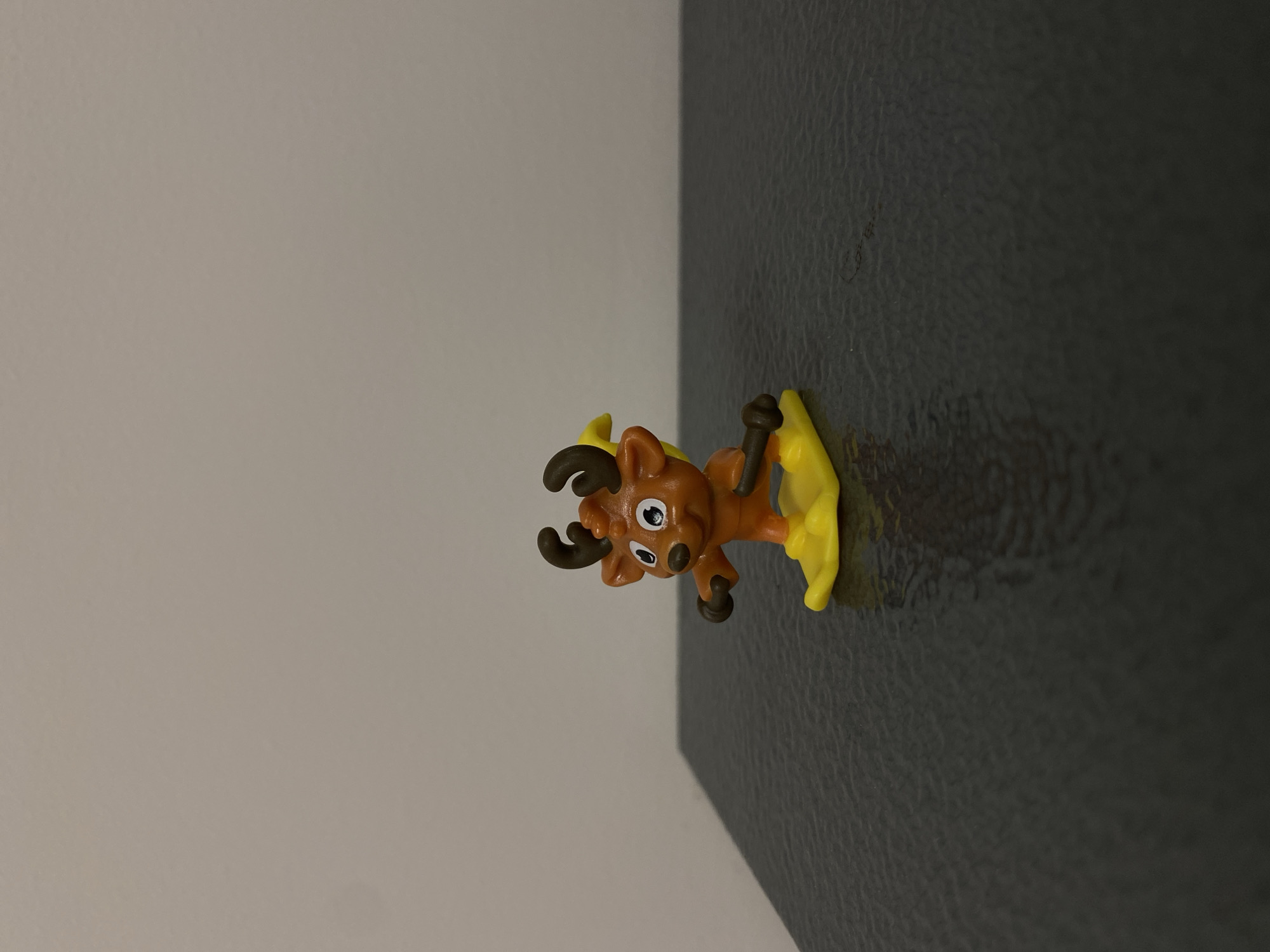 A close-up shot depicts a small, plastic toy reindeer. The toy is primarily orange, with brown antlers and yellow accents. It rests on a dark, textured surface, possibly a counter or shelf, which is contrasted by a light grey wall in the background. There is no contextual information to infer location in Votkinsk, Russia.