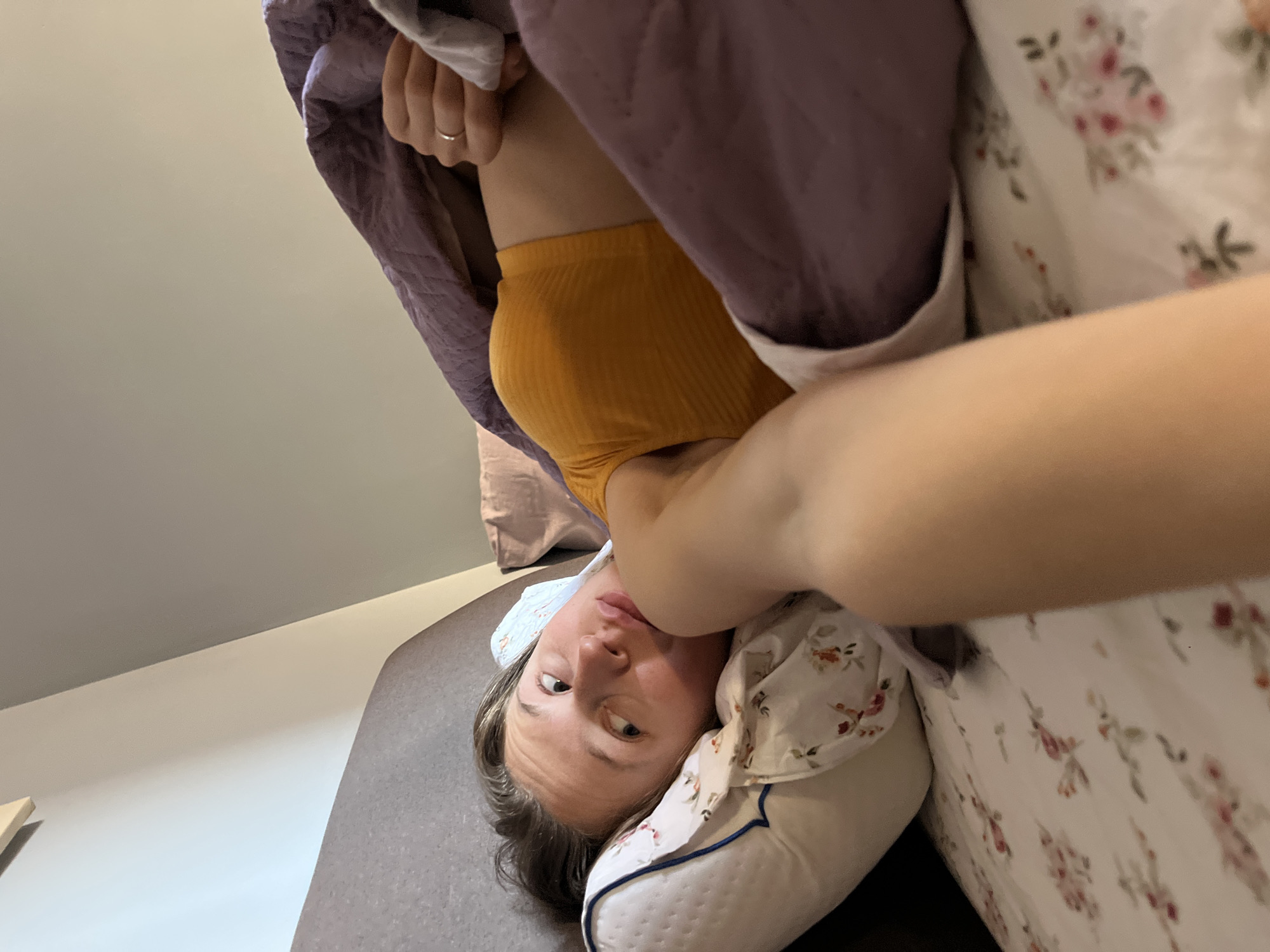 A woman is lying on a bed with a floral patterned pillow and duvet. She is wearing a yellow top and is partially covered by a purple duvet. She is looking at the camera. The location is likely indoors but the city of Bečići, Montenegro cannot be confirmed from the image.