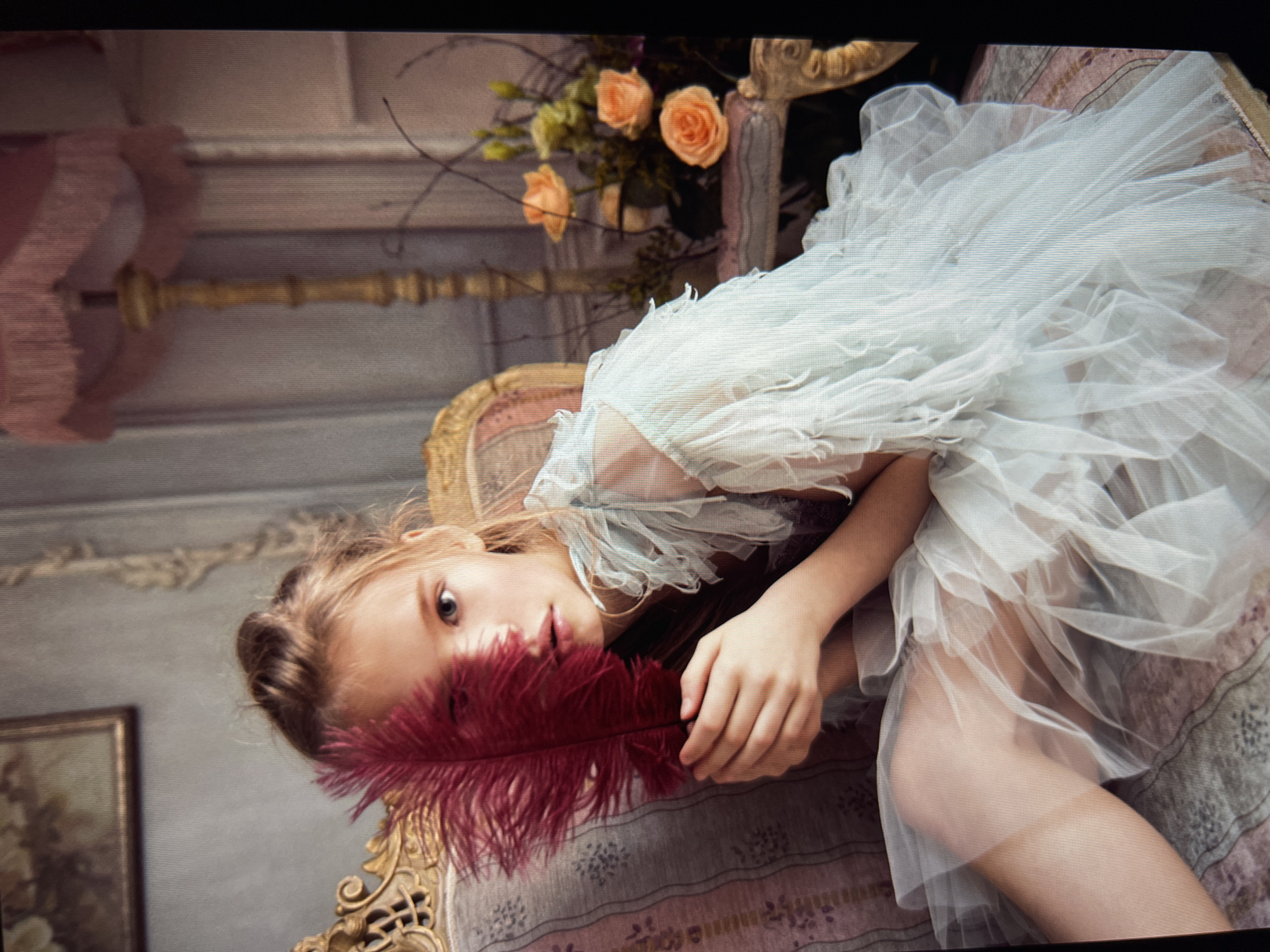 This is a portrait of a young girl lying down on a patterned surface, possibly a bed or couch, in what appears to be a dimly lit, ornate room. She is wearing a delicate, pale blue, ruffled dress. Her head is turned towards the camera, and she is holding a large, deep red feather in front of her face, obscuring her mouth and nose. Her eyes are wide and looking directly at the viewer.
In the background, there's a hint of a floral arrangement with peach-colored roses and greenery, suggesting a decorative element within the room. There are also glimpses of ornate furniture, possibly a chair with carved golden details and a pink upholstered headboard. A framed picture hangs on the wall behind her. The overall lighting is soft, with shadows casting on the girl's body and the surrounding elements. The scene evokes a sense of softness and perhaps a touch of melancholy or contemplation due to the girl's gaze and the feather's placement. There is no visible text in the image.