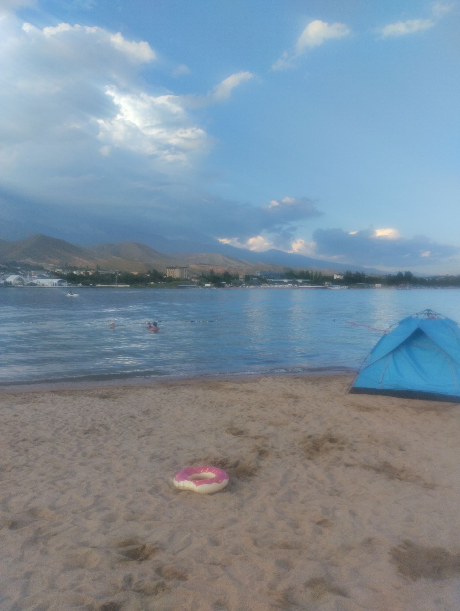 This image depicts a scenic view of a beach in Cholpon-Ata, Kyrgyzstan, with the Issyk-Kul lake in the foreground and mountains in the background. A blue tent is pitched on the sandy beach to the right of the frame. In the water, several people are swimming, and a jet ski can be seen in the distance. A pink inflatable donut-shaped float rests on the sand in the foreground. The sky is partly cloudy with a mix of sunlight and shadows, suggesting it is either early morning or late afternoon. The overall atmosphere is one of a tranquil summer day at the lake.