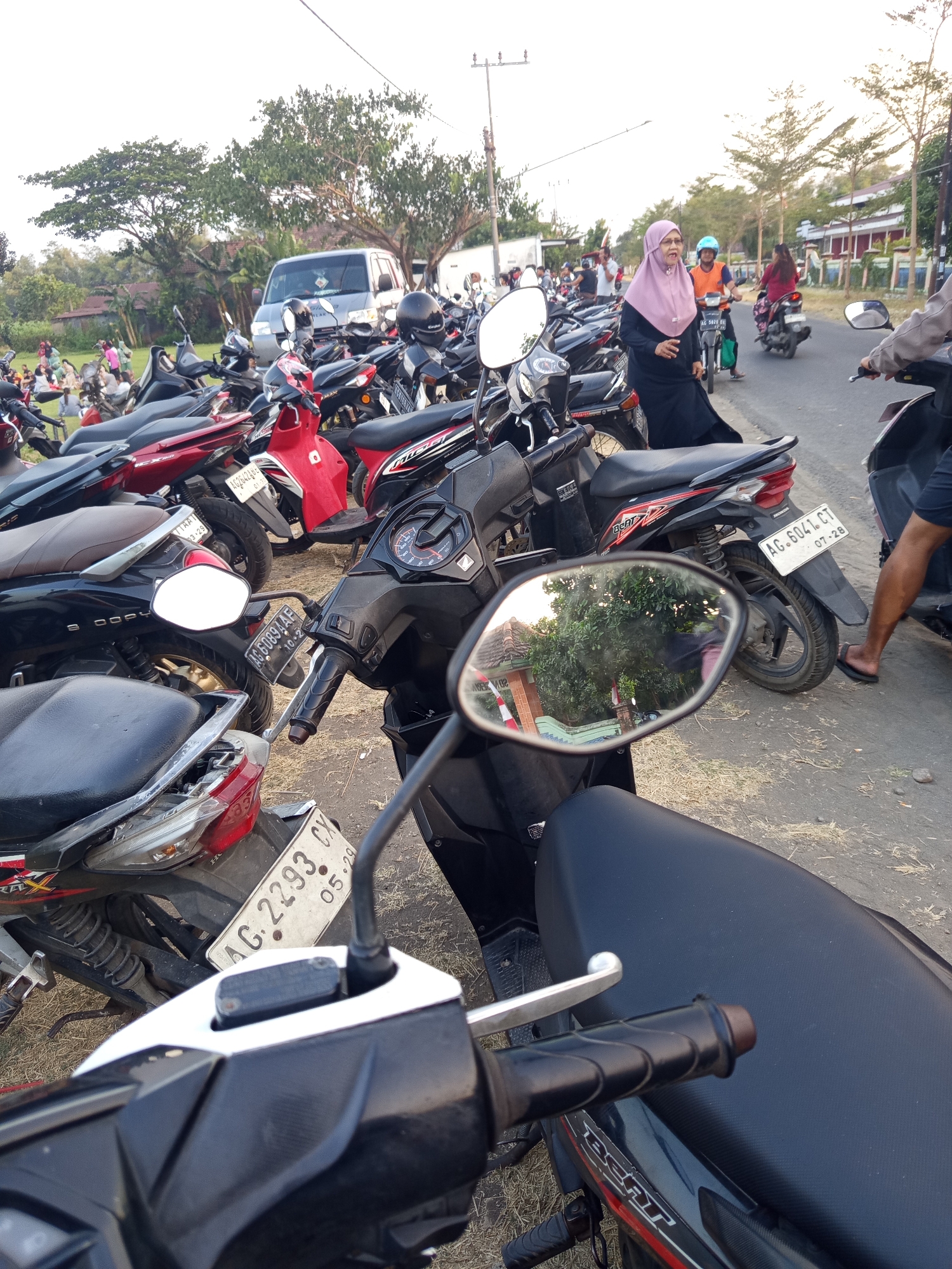 The image captures a densely packed outdoor parking area for motorcycles in Mrican, Indonesia. A significant number of scooters and motorcycles are parked in rows on an unpaved surface, with some on the asphalt road. Several people are visible, some walking amongst the parked vehicles and others on motorcycles. One woman wearing a pink hijab and dark clothing is walking in the foreground, looking towards the camera, while another person in an orange shirt and blue helmet rides a motorcycle in the background. A silver van is parked amongst the motorcycles in the distance. The environment appears to be semi-rural or suburban, with trees and buildings visible in the background under a bright sky. The foreground is dominated by the handlebars, mirrors, and seats of several motorcycles, suggesting the image was taken from the perspective of someone sitting on or standing near a parked motorcycle. License plates are visible on some of the motorcycles, with inscriptions like "AG. 6041 CT" and "AG. 2293 CX." The overall scene depicts a busy gathering or event where many individuals have arrived by motorcycle.