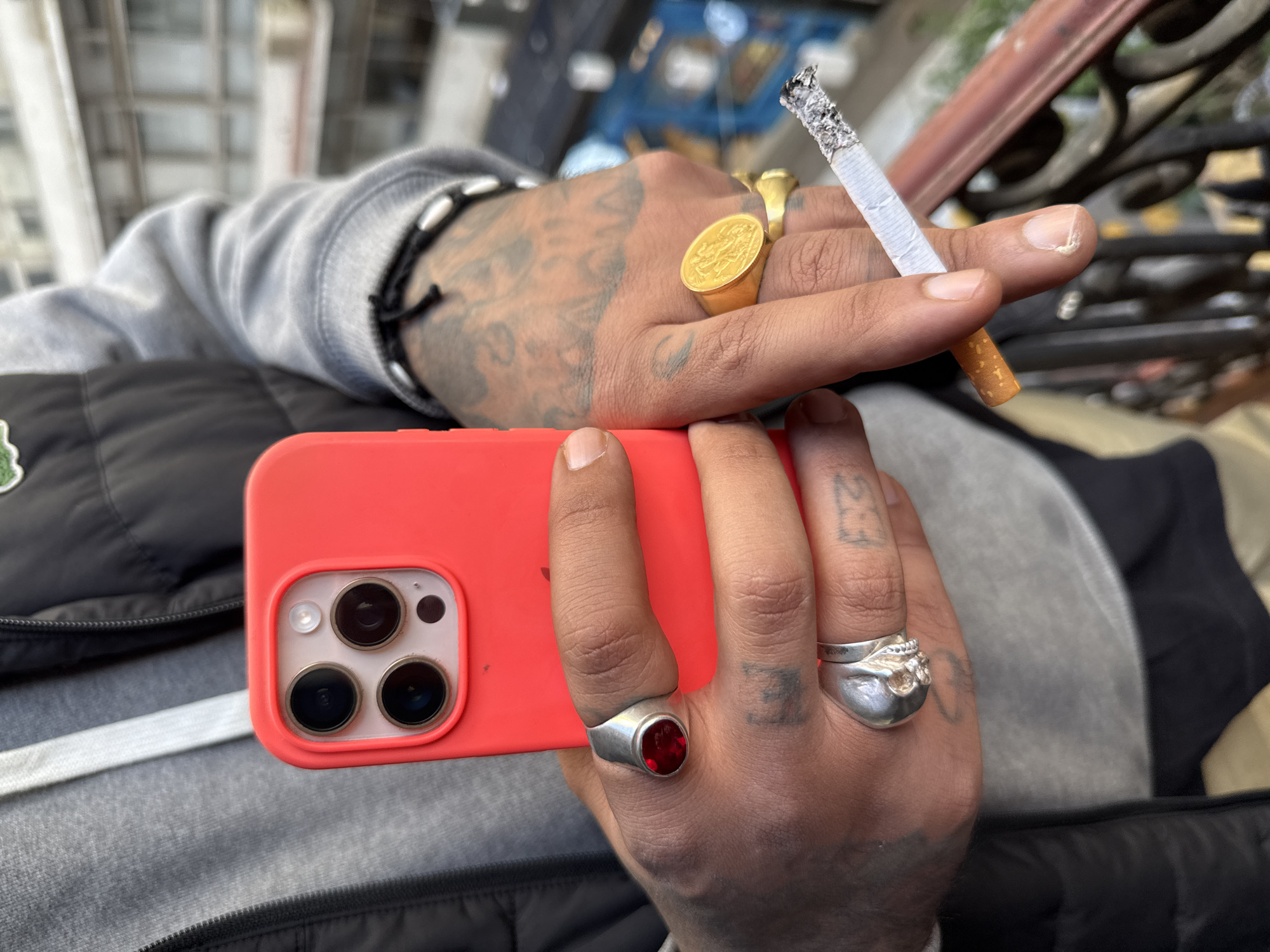 A person holds a red cell phone with a triple camera lens in their left hand, while a lit cigarette is held between the thumb and forefinger of the right hand. The hands are adorned with several rings, including a large gold ring, a silver ring with a red stone, and a skull-shaped ring. The person is wearing a grey hooded sweatshirt and a black vest. The background appears to be an outdoor urban setting with wrought iron details, suggesting a location in a city; however, pinpointing the exact location in Buenos Aires, Argentina, is not possible from the image alone.