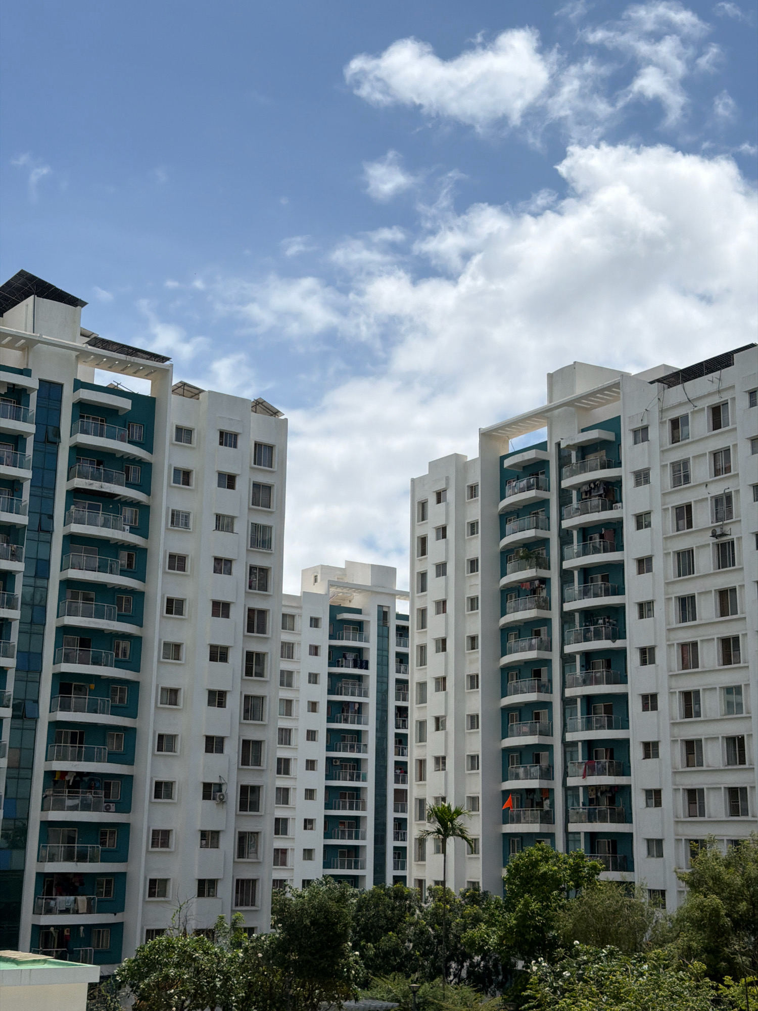 The image shows a residential complex in Pune, India, on a day with a blue sky and scattered clouds. The complex consists of several tall, white apartment buildings with teal accents and balconies. The buildings have many windows, and some balconies have laundry hanging. Solar panels are visible on the roofs of some buildings. Lush green trees and foliage surround the base of the buildings. A single palm tree stands in front of one of the buildings. There are no people visible in the image. The scene appears to be a typical residential area in a modern Indian city.